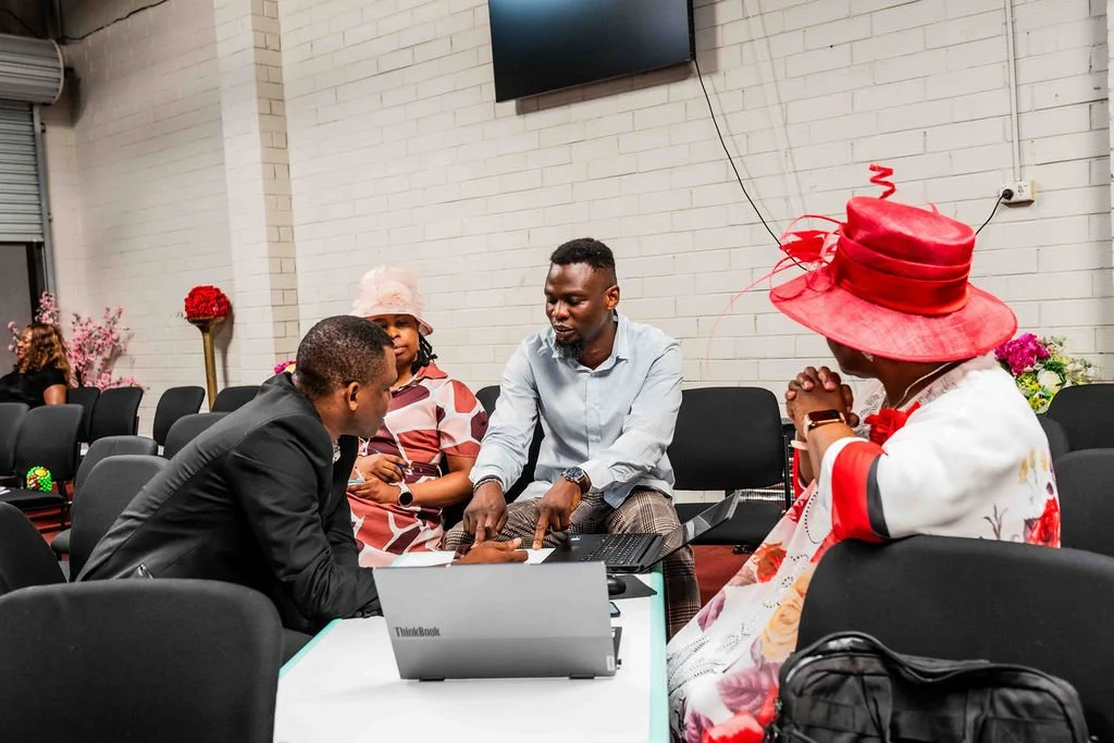 Group of adults meeting around laptops and paperwork in a church or community room, with a woman wearing a bright red hat.