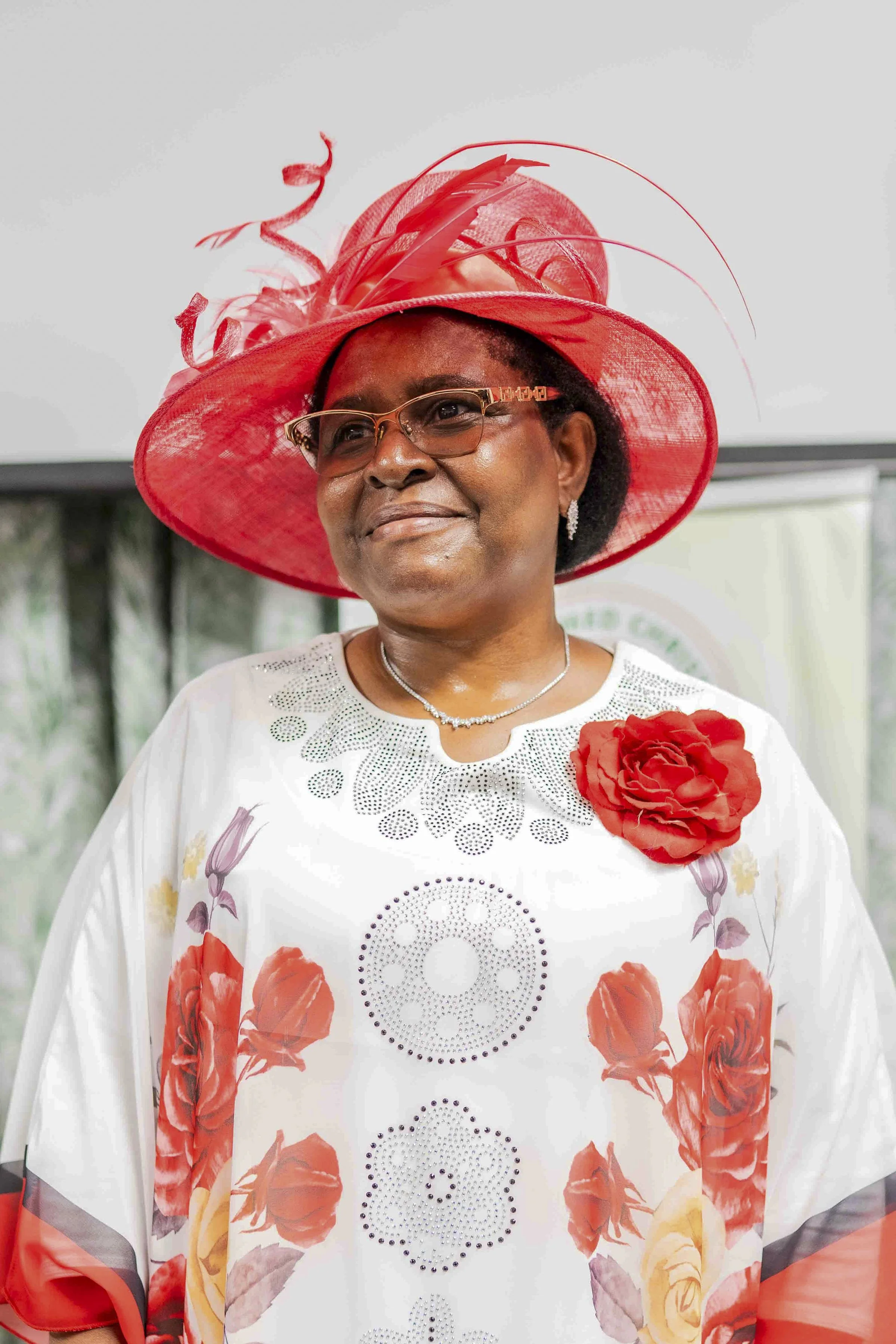 Portrait of an older Black woman wearing glasses, a large red fascinator, and a white floral dress with a red rose corsage