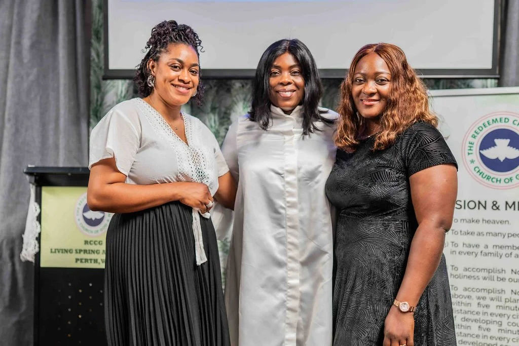 Three smiling women posing together at a church event in front of a presentation screen and church signage.