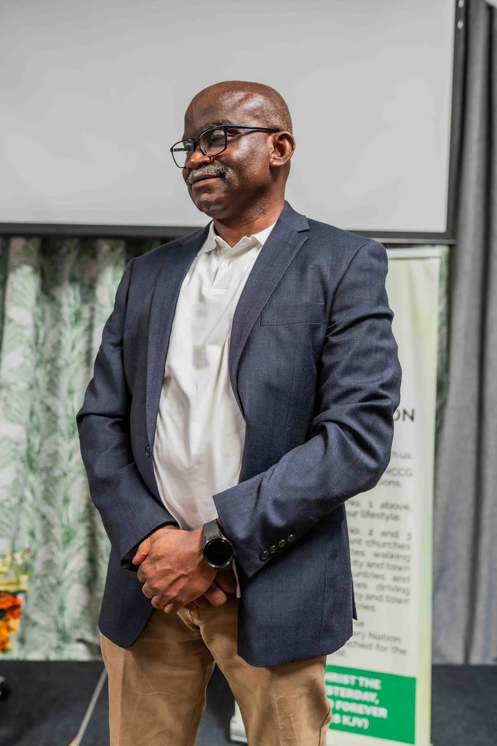 Man wearing glasses, a blue blazer, white shirt, and smartwatch standing in front of a presentation screen.