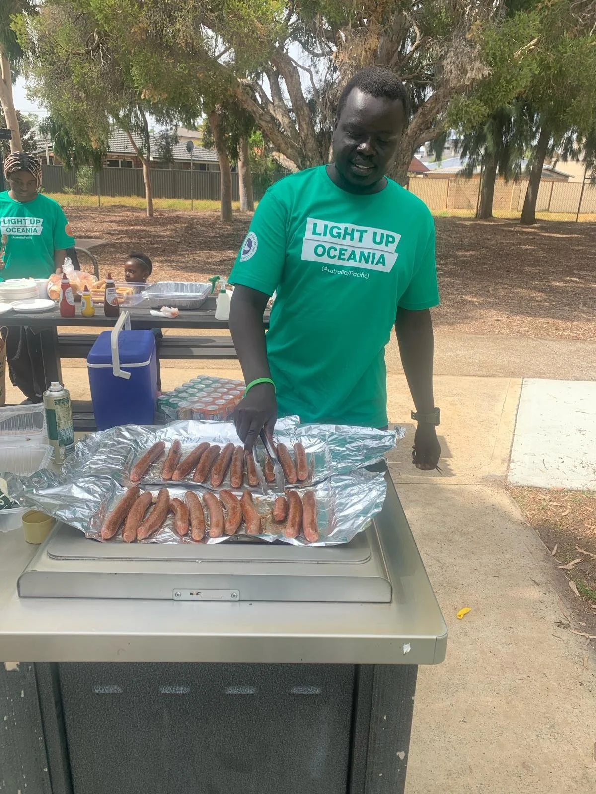 Man grilling sausages on an outdoor barbecue table during a community event.