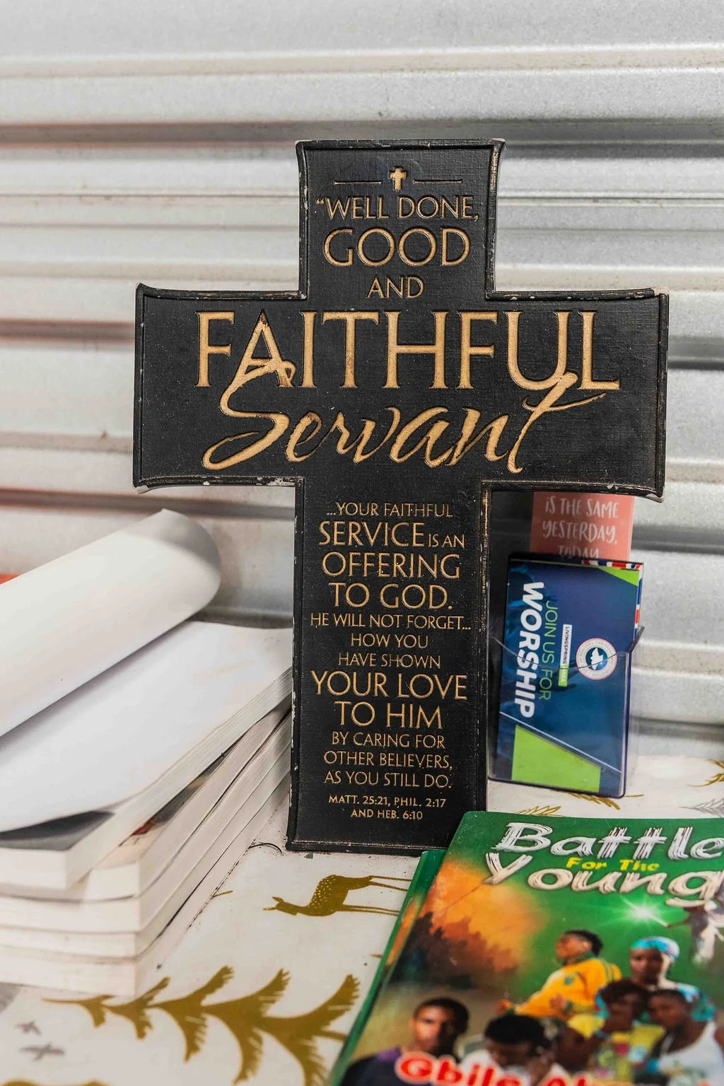 Black and gold Christian cross plaque reading “Well done, good and faithful servant,” displayed beside books and a worship booklet.