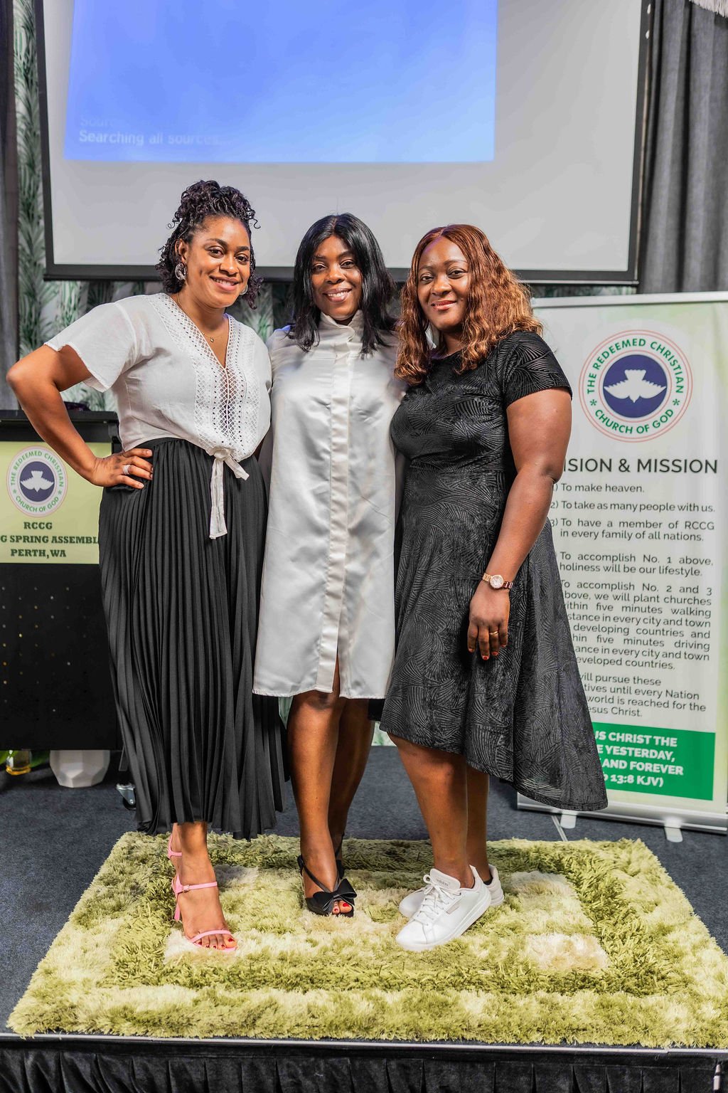 Three women posing together at a church event in front of a projected screen and Redeemed Christian Church of God banner