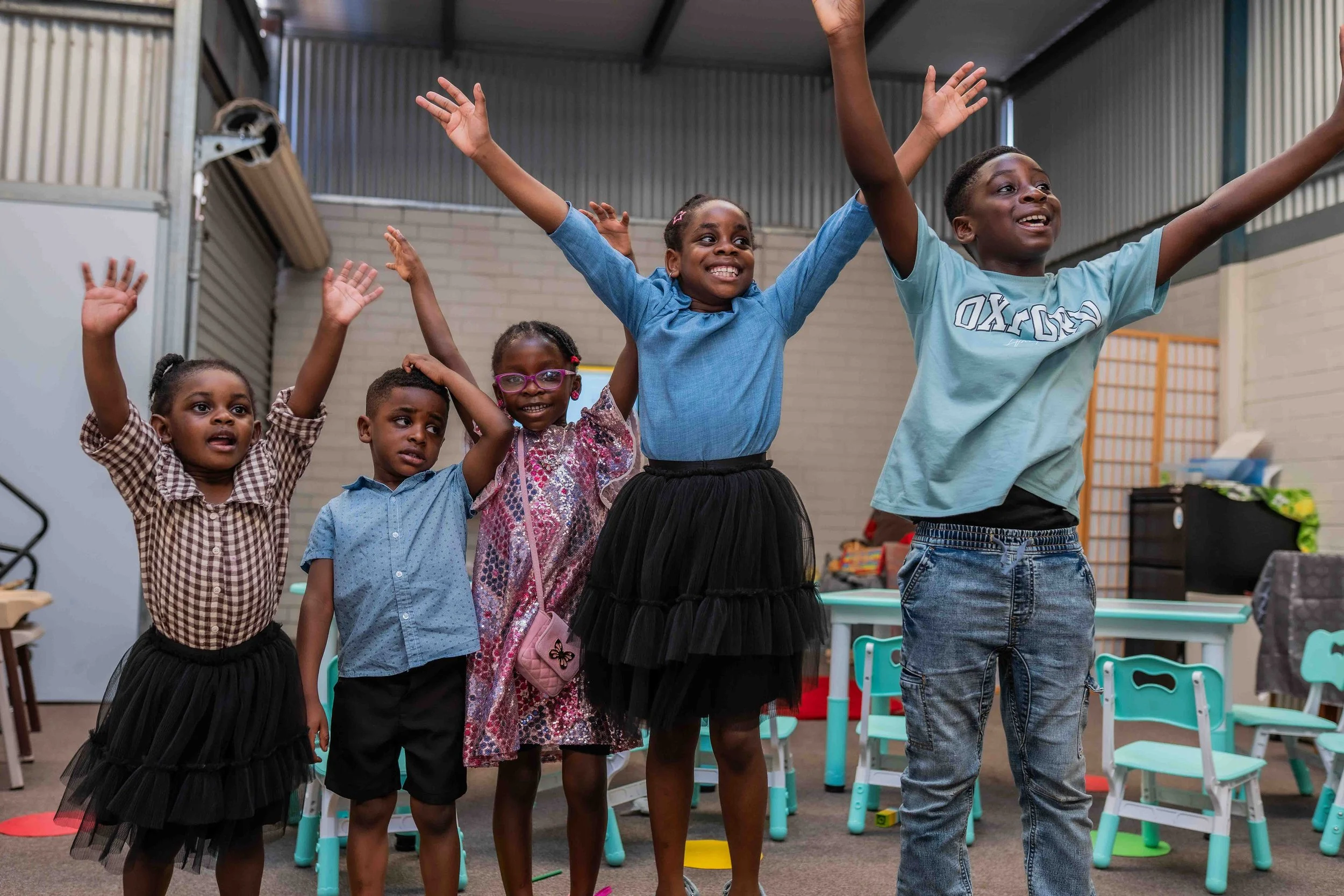 Five children smiling and raising their hands in a colorful indoor classroom or playroom