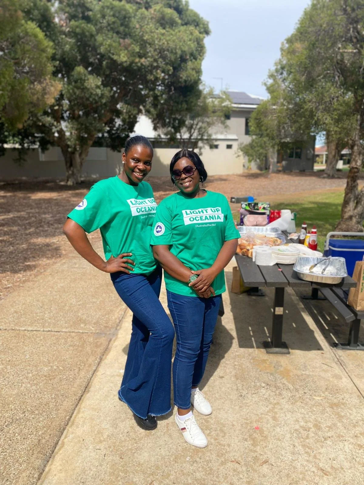 Two women wearing green t-shirts with 'Light Up Oceania' written on them, standing outdoors near a picnic table with food and supplies, smiling at the camera.