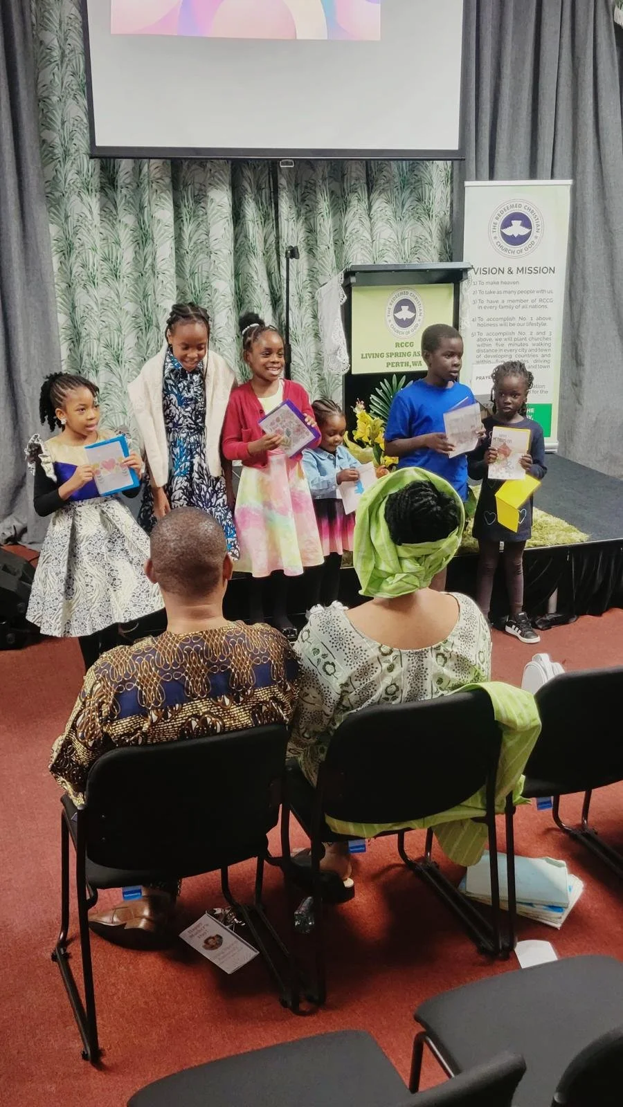 Children standing on stage during a presentation at a church event, some holding books or papers, with an audience watching in front.