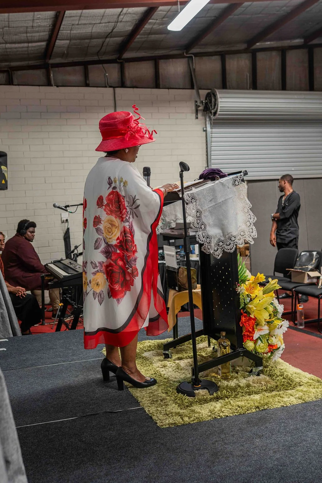Woman in a red floral dress and hat speaking at a decorated podium while a pianist performs in the background.