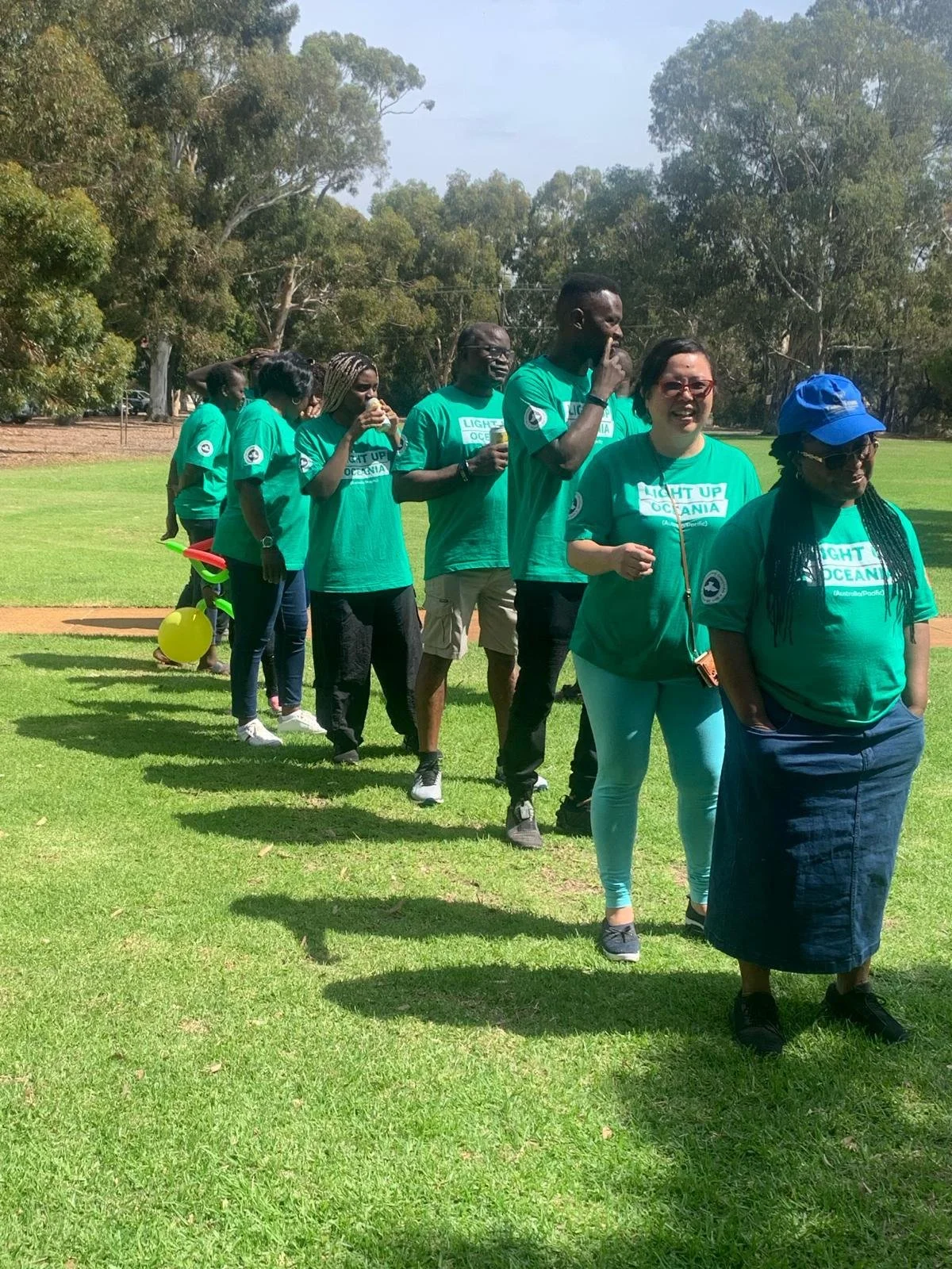 A group of people standing in a line on a grassy field, wearing matching teal shirts with the message 'Light Up Oceania'.