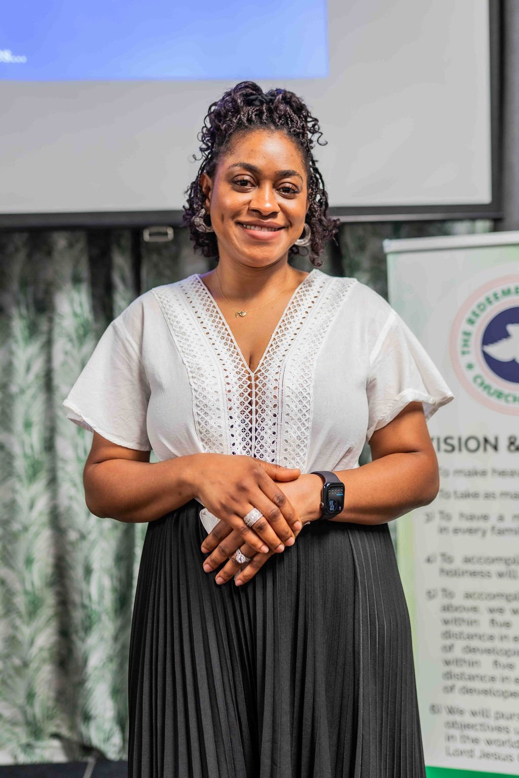Smiling woman with curly hair standing in front of a presentation screen, wearing a white blouse and black pleated skirt