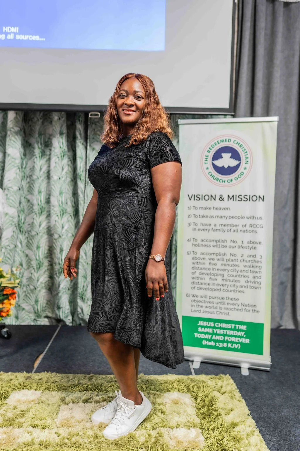 Smiling woman in a black dress and white sneakers standing beside a Redeemed Christian Church of God vision and mission banner.
