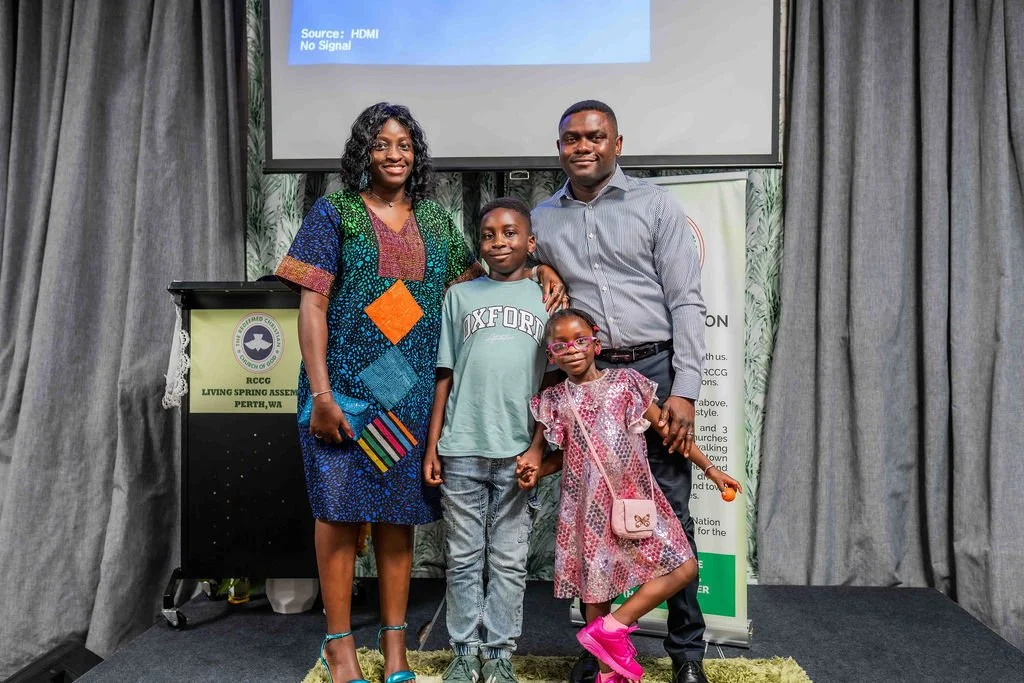 Family of four posing onstage beside a podium and projection screen at an assembly event