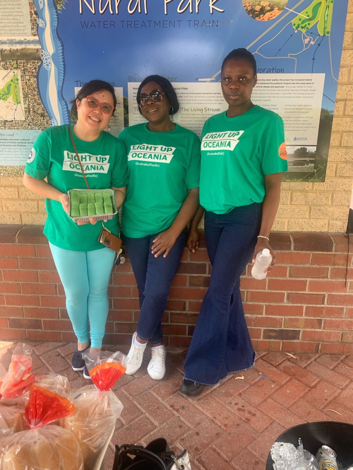 Three women standing outdoors in front of a blue informational board, wearing green t-shirts with 'Light Up Oceania' printed on them, holding a tray of green pastries and a water bottle, and there are plastic bags and a bucket with ice in the foregro