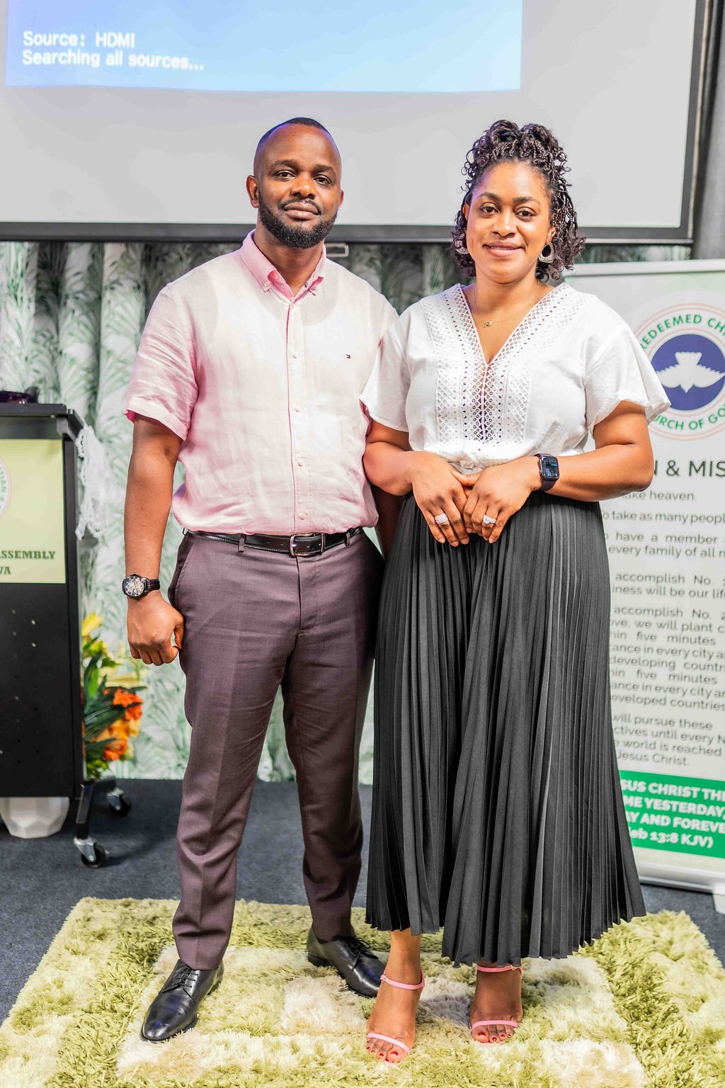 A man and woman pose together at a church event in front of a projector screen and church banner.