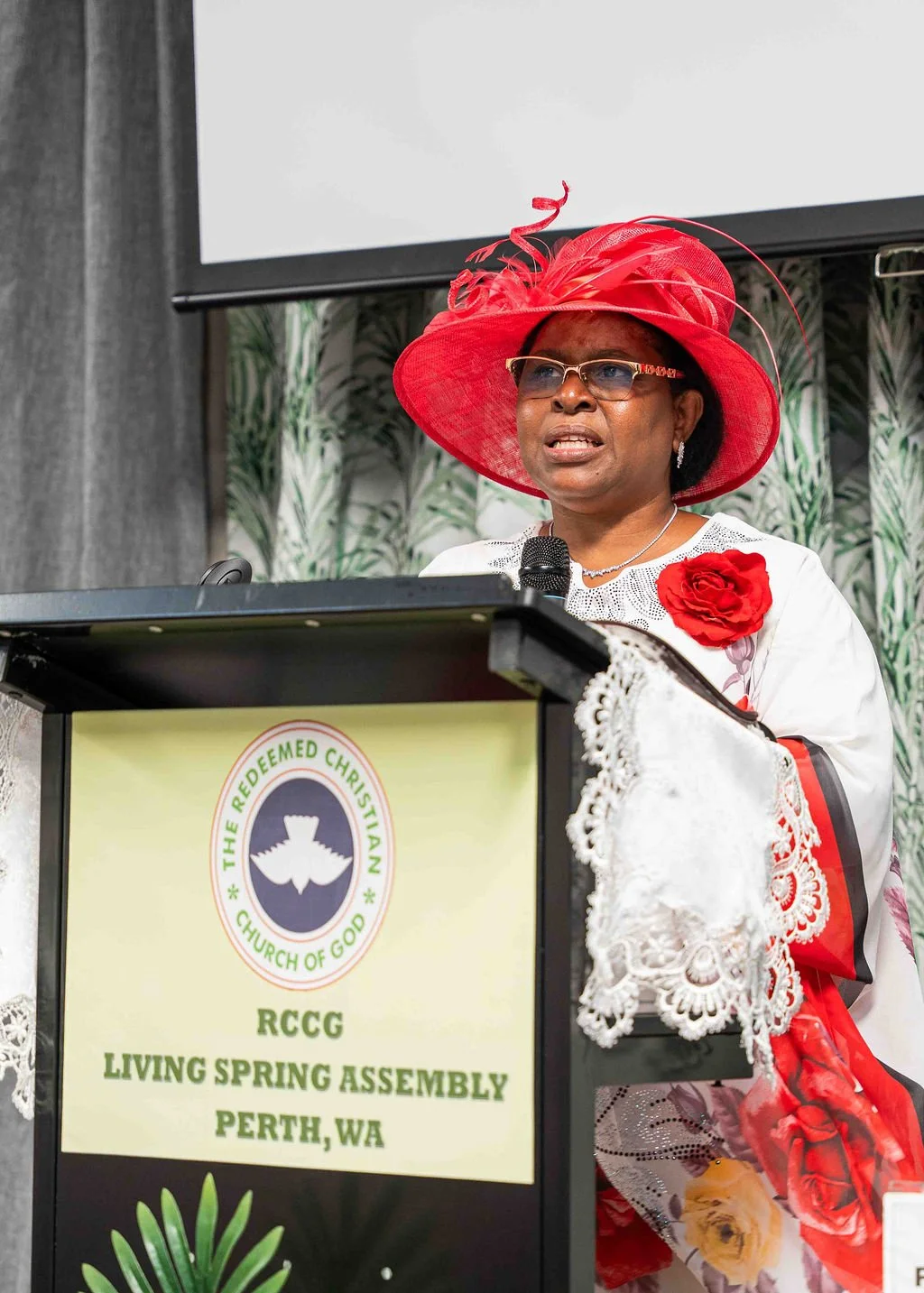 Woman in a red hat and white floral outfit speaking at a podium labeled RCCG Living Spring Assembly Perth, WA.