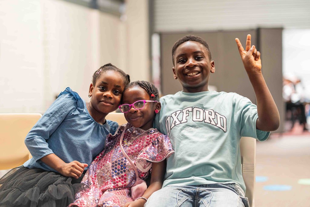 Three smiling Black children pose together indoors, with the boy wearing an Oxford shirt and making a peace sign.