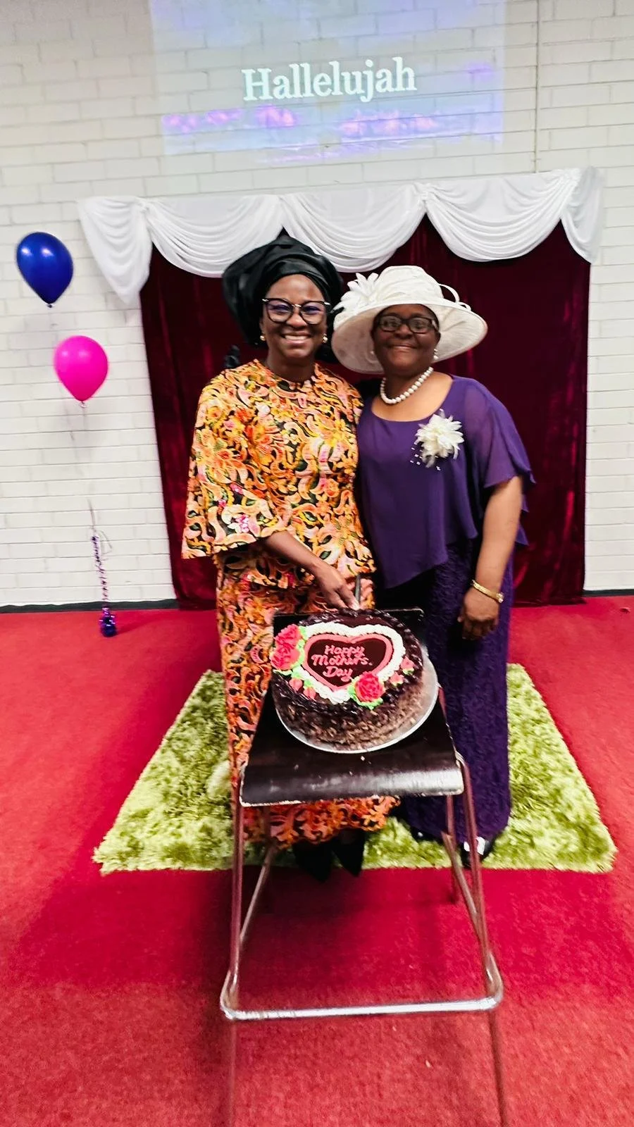Two smiling women, one with a large black headwrap and the other with a white hat, standing next to a chocolate cake that reads 'Happy Mother's Day' in a decorated room with balloons and a red carpet.