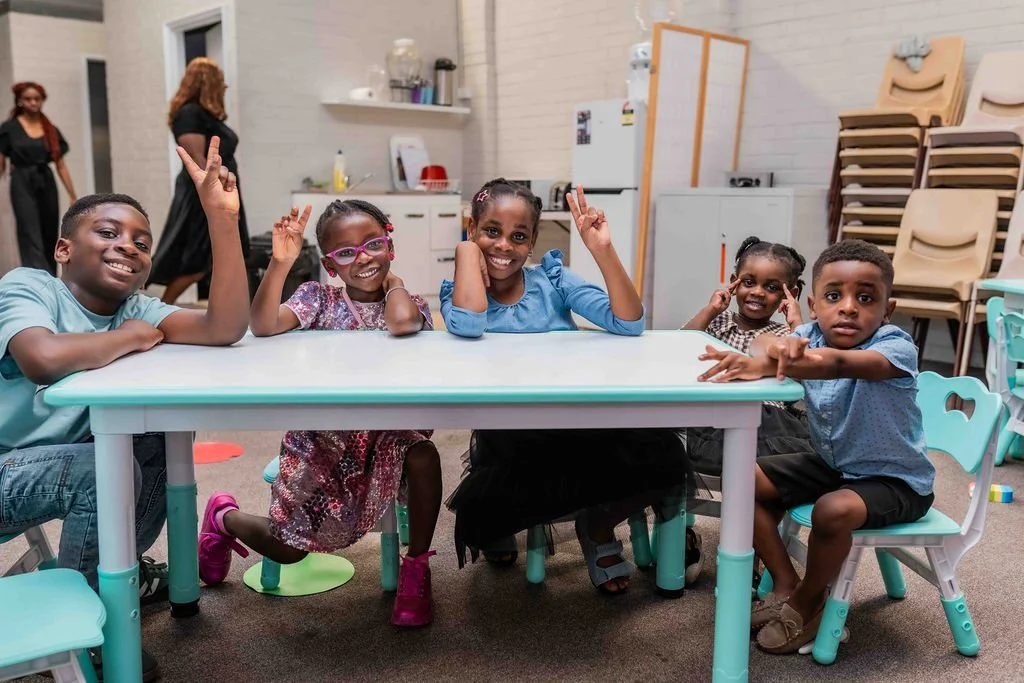 Five smiling Black children posing around a classroom table, with several raising peace signs.
