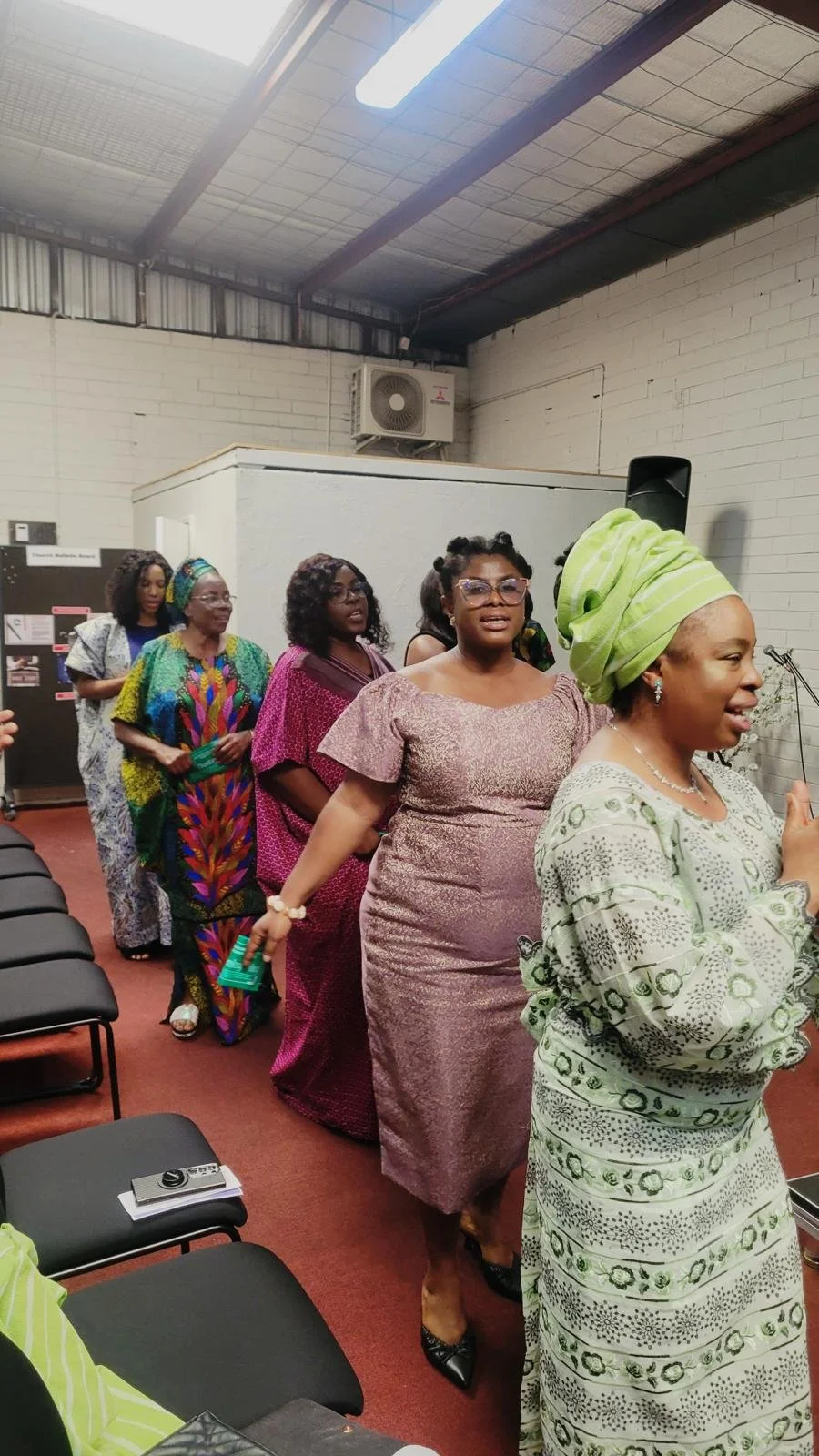 A group of women standing in a line, wearing colorful traditional African dresses, in an indoor setting with ceiling lights and chairs.