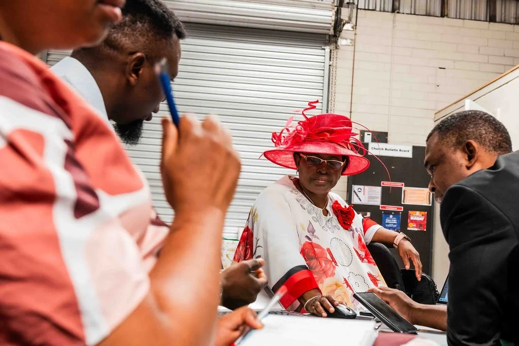 Older woman in a bright red hat meeting with two men at a desk in an industrial office.