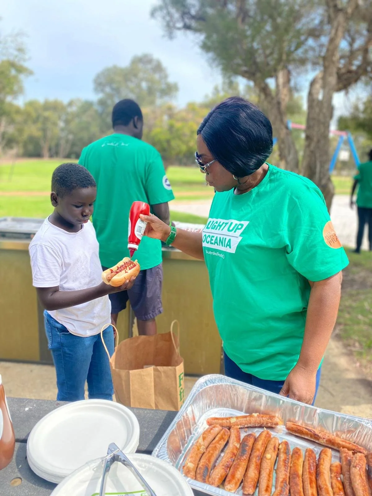 A woman in a green T-shirt applying ketchup to a hot dog held by a young boy, with a table of cooked hot dogs and supplies outdoors in a park.
