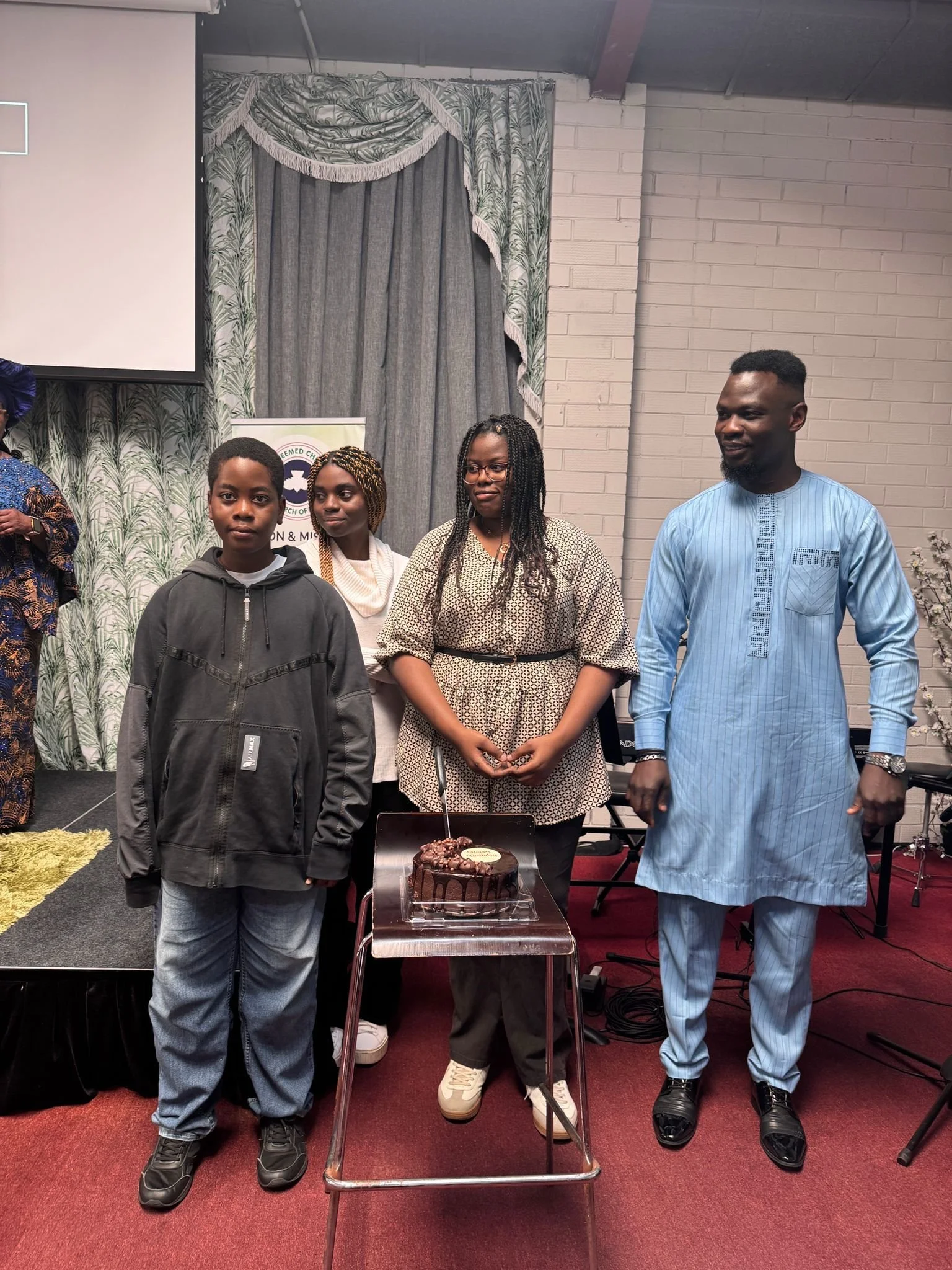 A group of four people standing indoors in front of a table with a chocolate cake. The setting appears to be a celebration or event, with a decorative curtain and banner in the background.