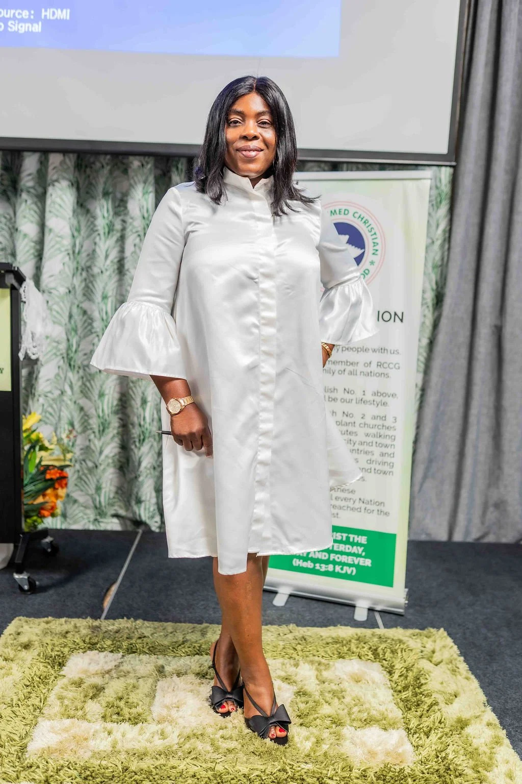 Woman in a white dress standing at a church event beside a presentation screen and banner