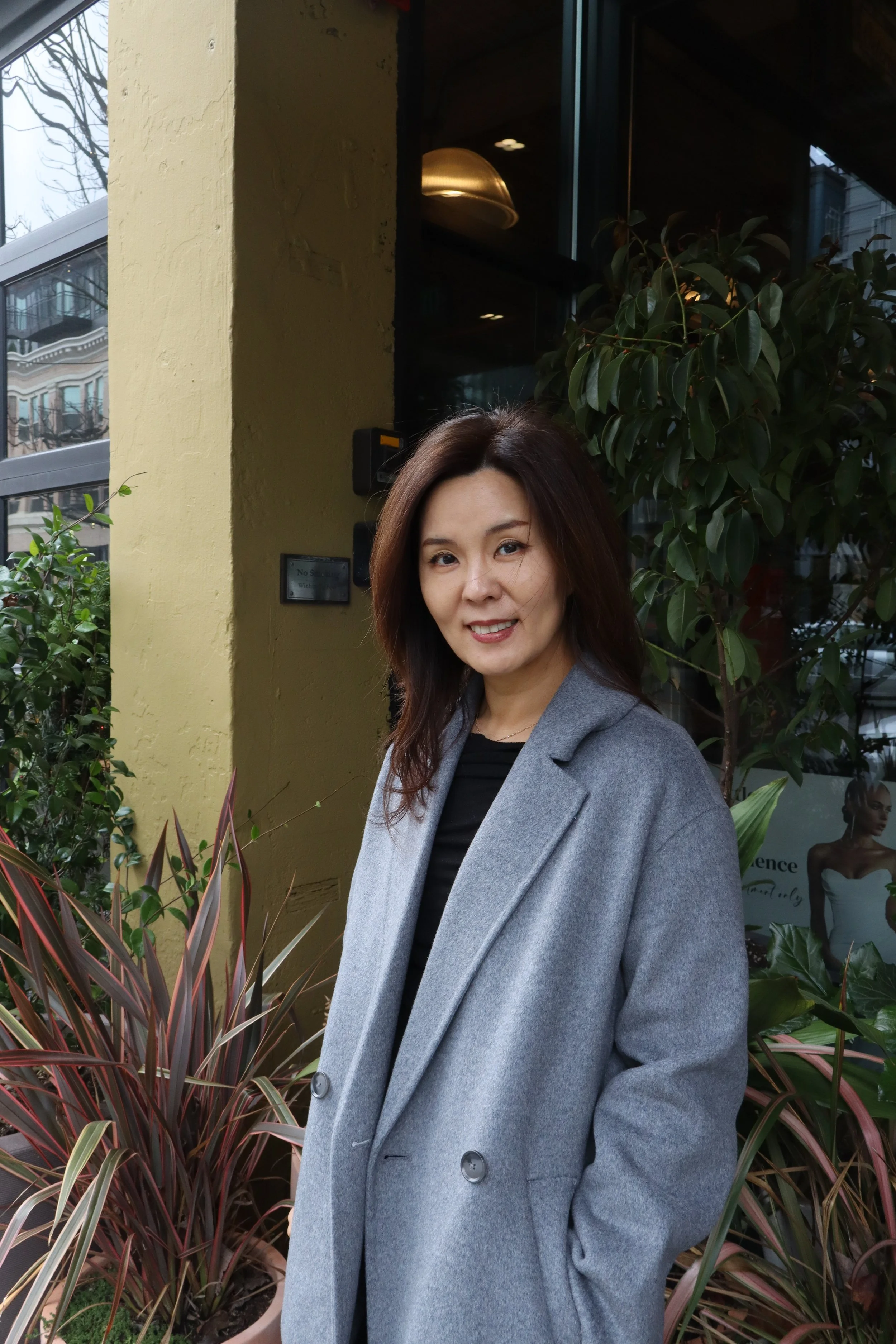 A woman with shoulder-length brown hair smiling, wearing a black turtleneck, gray cardigan, and black skirt, standing with her arms crossed in front of a stone wall with a modern clock.