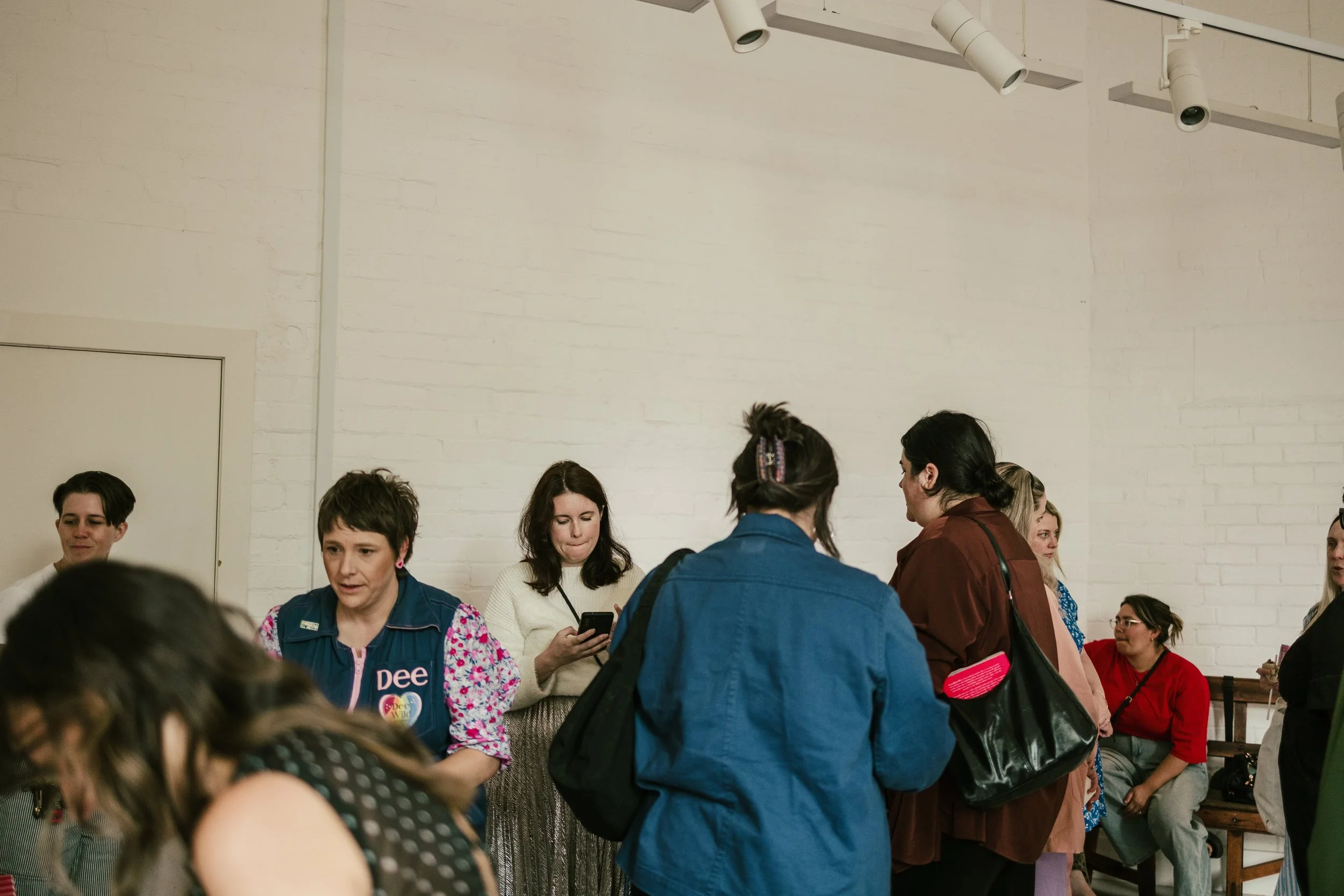 Group of women at a Bento and Bubbles Celebration event talking and looking at their phones inside a room with white brick walls.