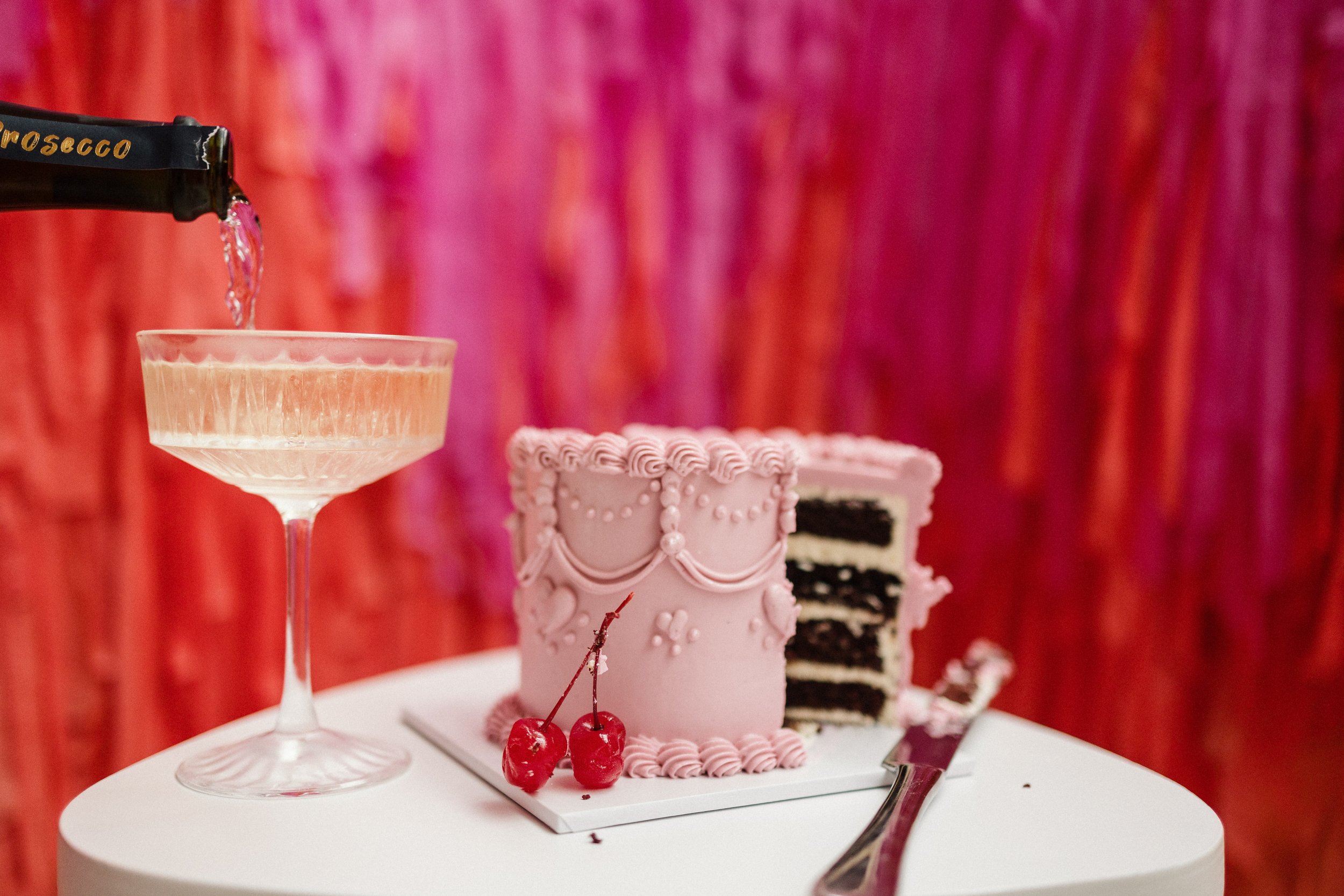 A pink frosted cake with decorative icing and two cherries on top, partly sliced to reveal black and white layers, next to a cocktail glass with a pink drink, and a silver knife, on a white table with a red and pink backdrop.