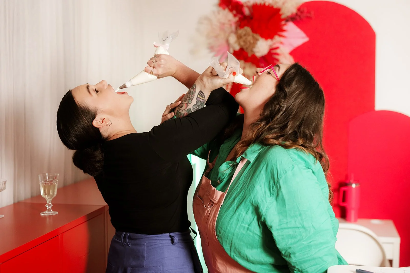 Two women engage in a playful moment as one pipes buttercream into the other's mouth at a celebration, against a decorated red backdrop.