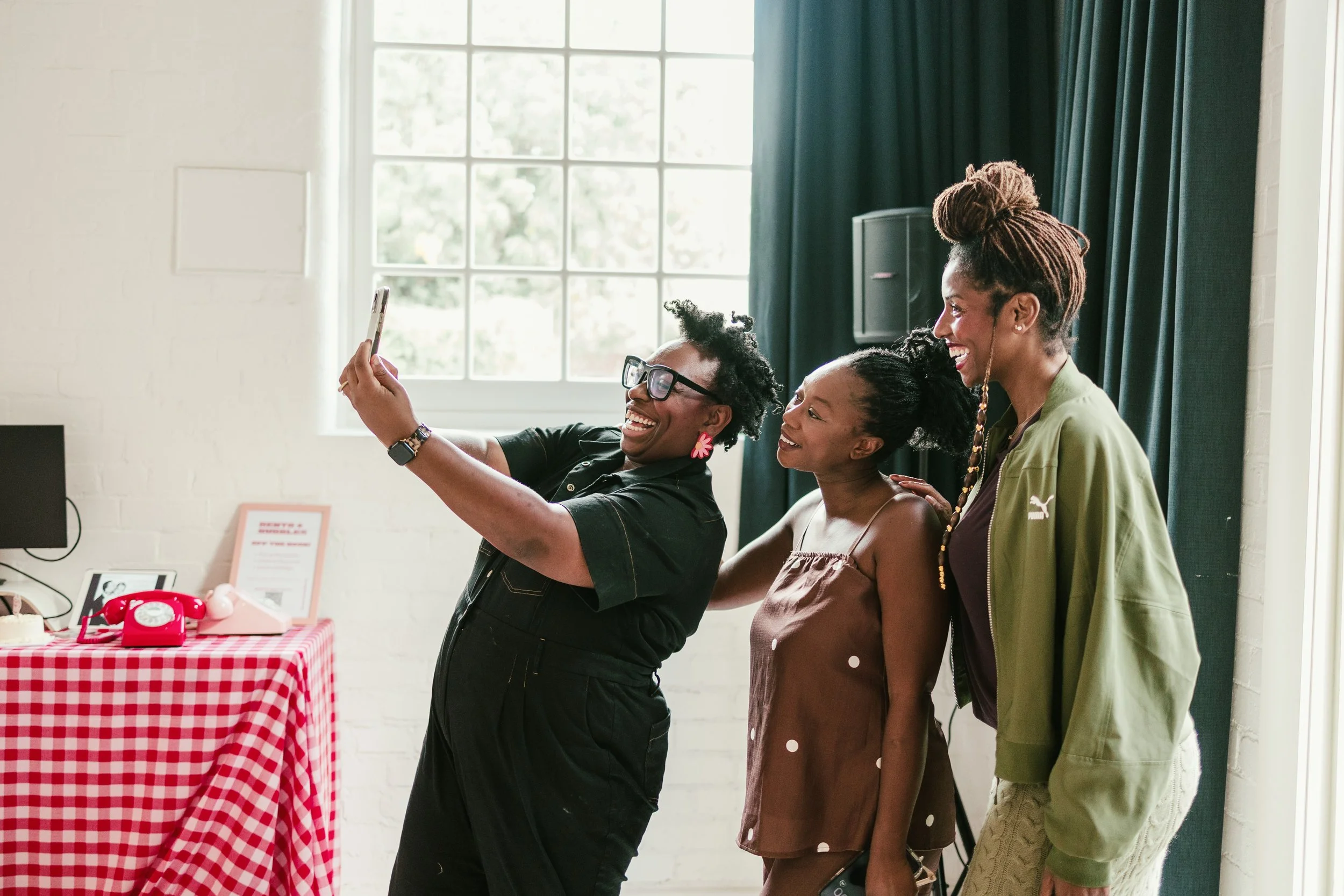 Three women taking a selfie indoors with a window and dark curtains in the background. One woman is holding a phone, and they are all smiling and posing.