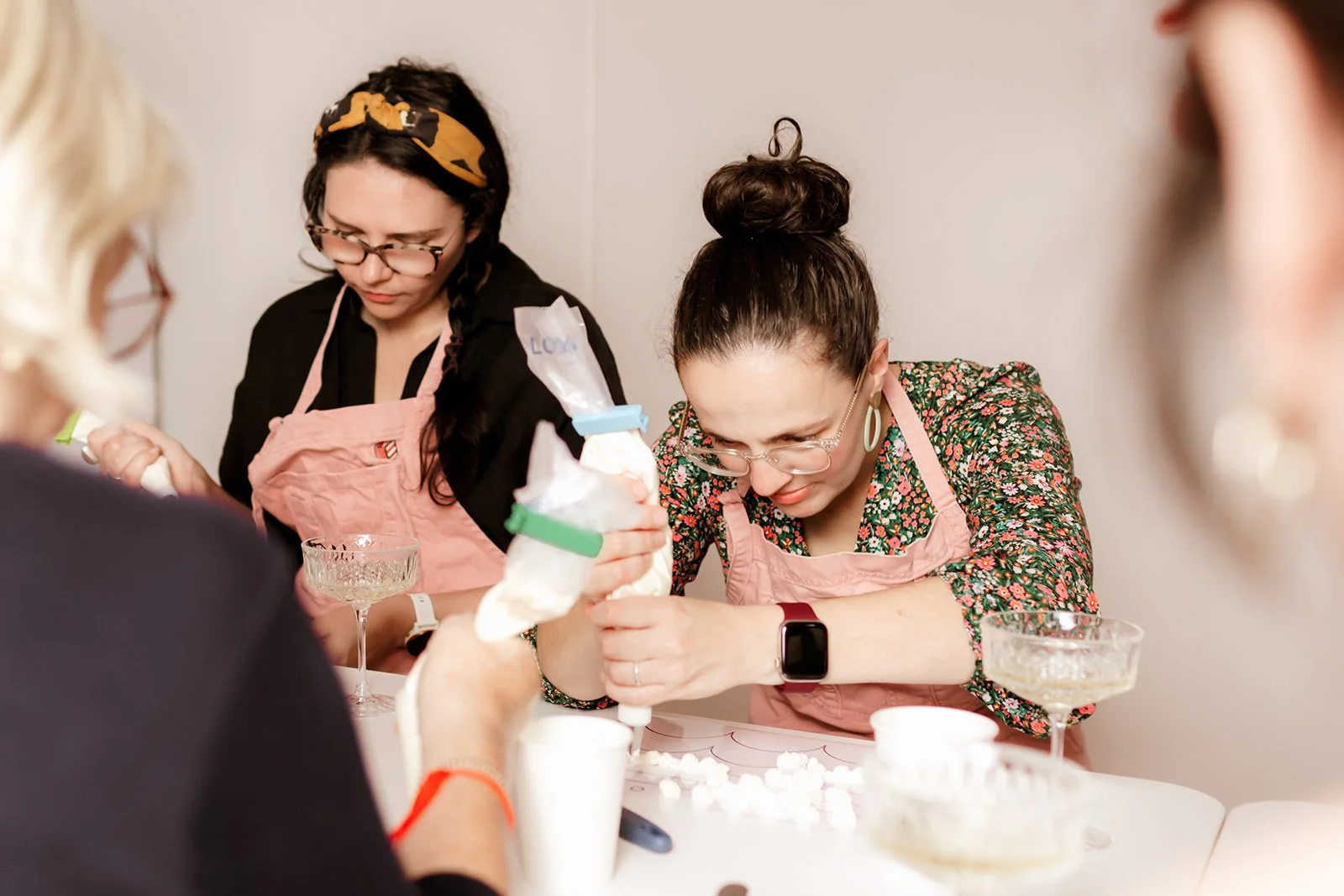 Women in aprons and glasses working together at a table, filling bags with buttercream for a baking project.