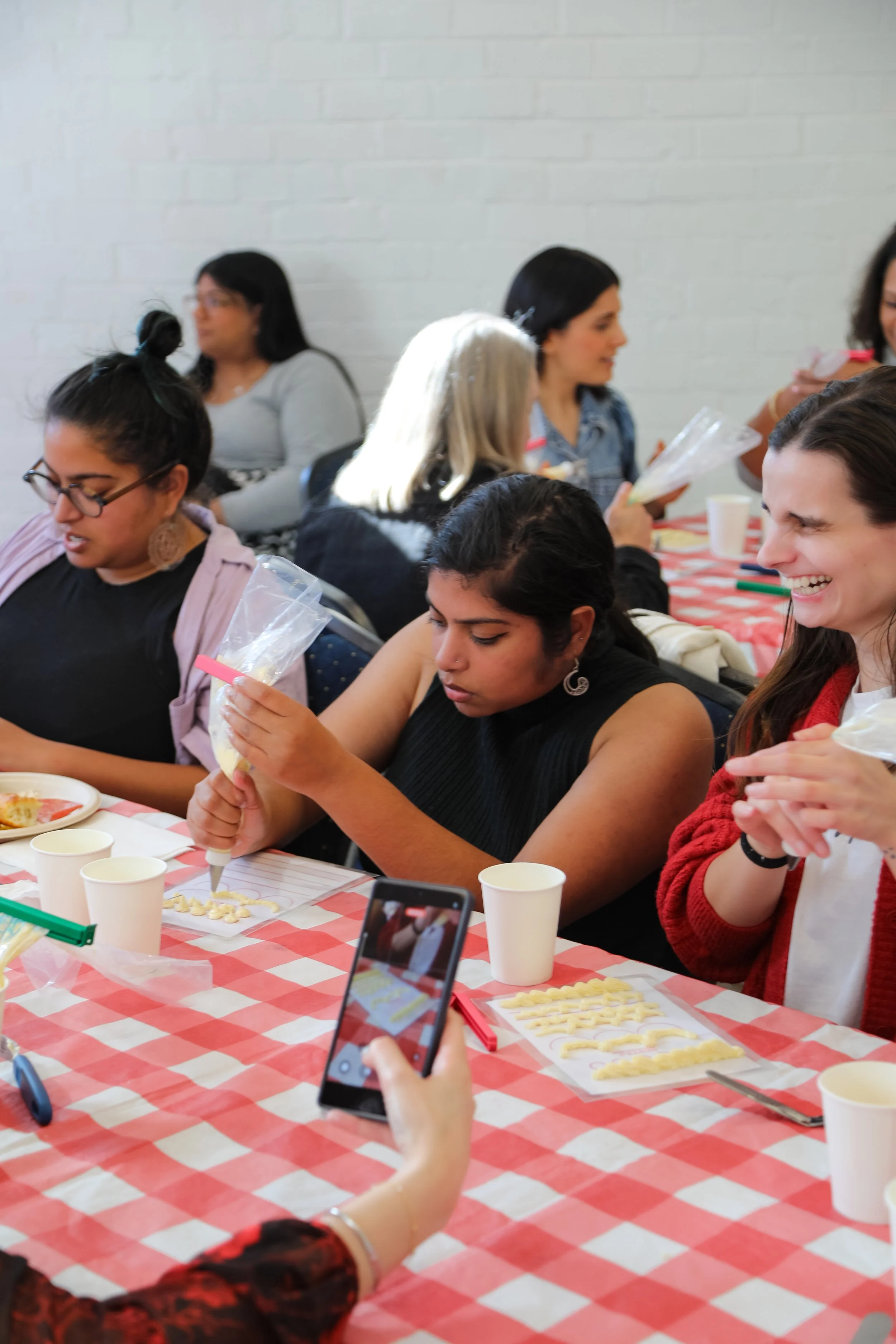 Group of women sitting at a table with a red and white checkered tablecloth, decorating cookies using piping bags, and one woman is taking a photo with her phone.