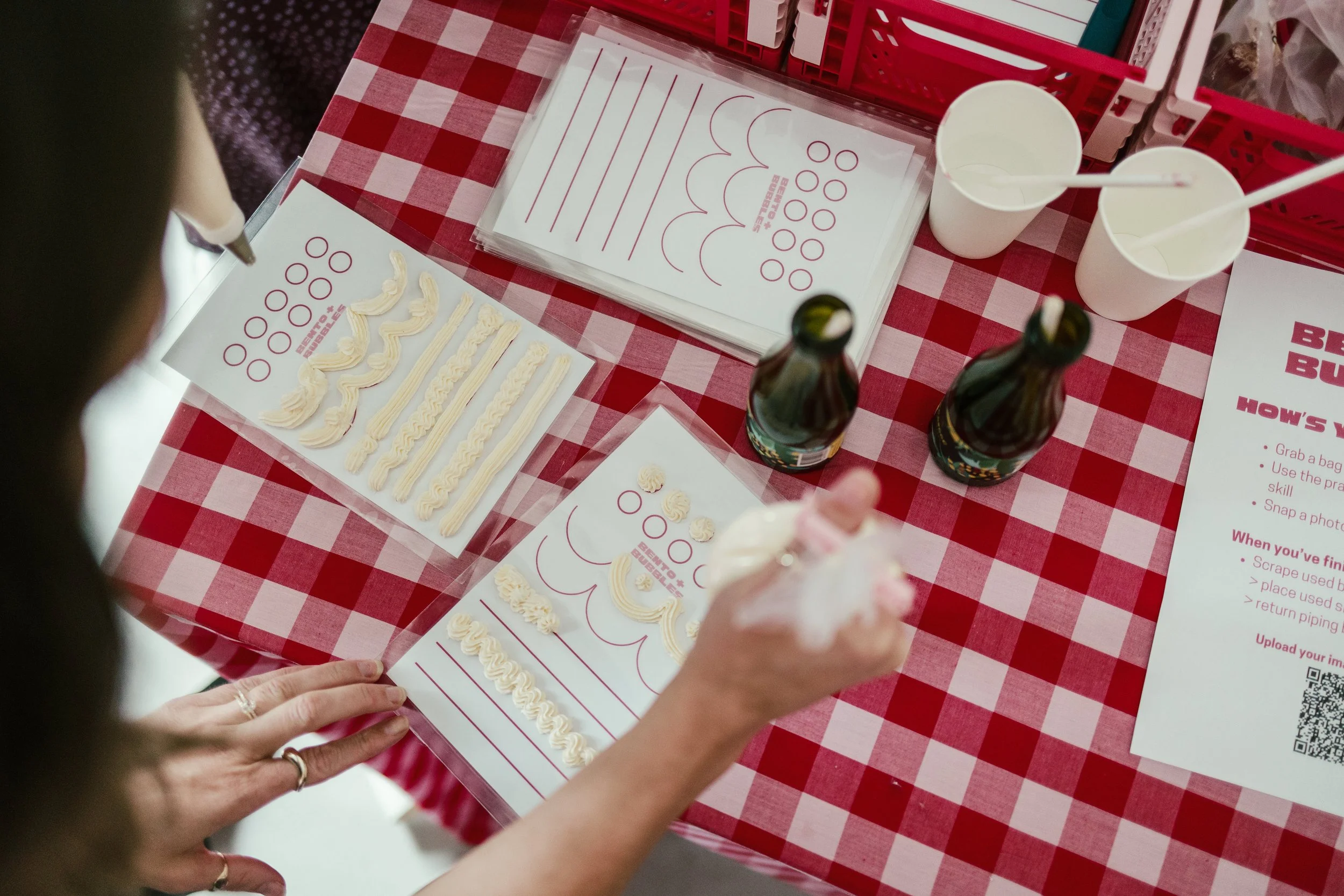 A person piping white frosting onto a sheet with various shapes on it at a table with a red and white checkered tablecloth. There are some empty cups, small bottles, and information sheets on the table.