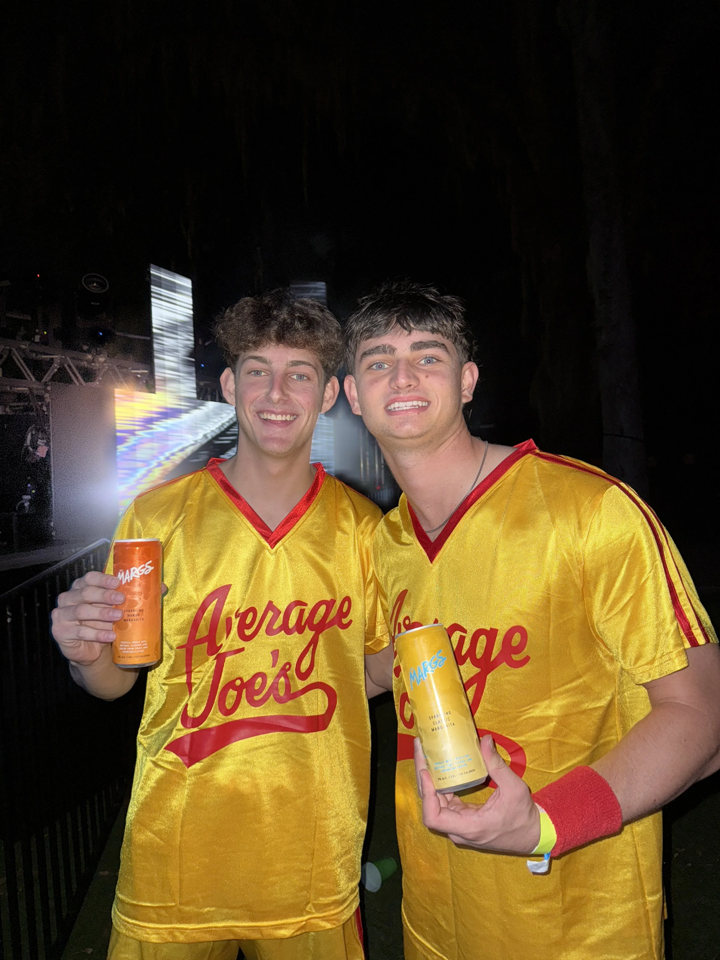 Two young men in yellow jerseys with red lettering that says "Average Joe's" are smiling and holding cans of Marges drinks at an outdoor event or concert at night, with stage lighting in the background.