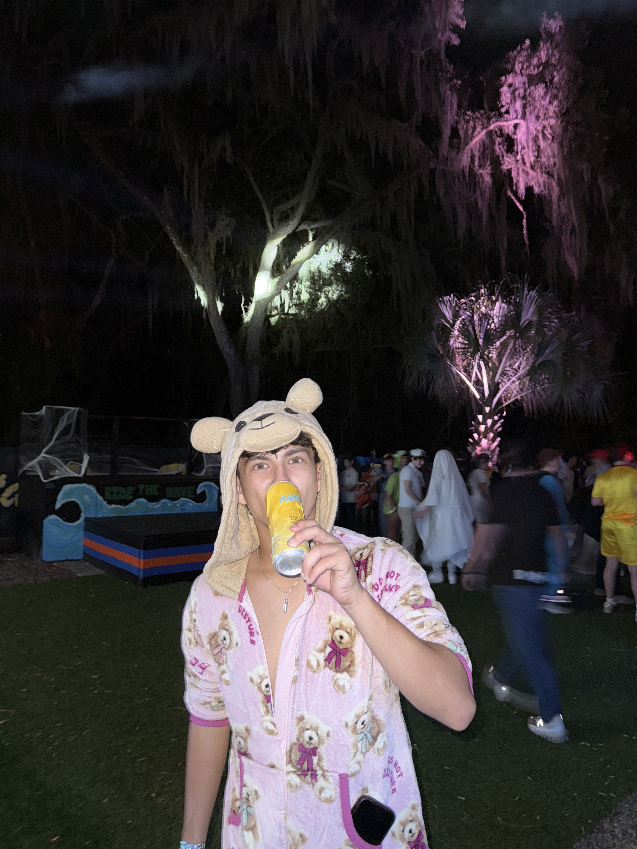 A person wearing a teddy bear hooded towel drinks from a can at a nighttime outdoor event with trees, colorful lights, and a crowd in the background.