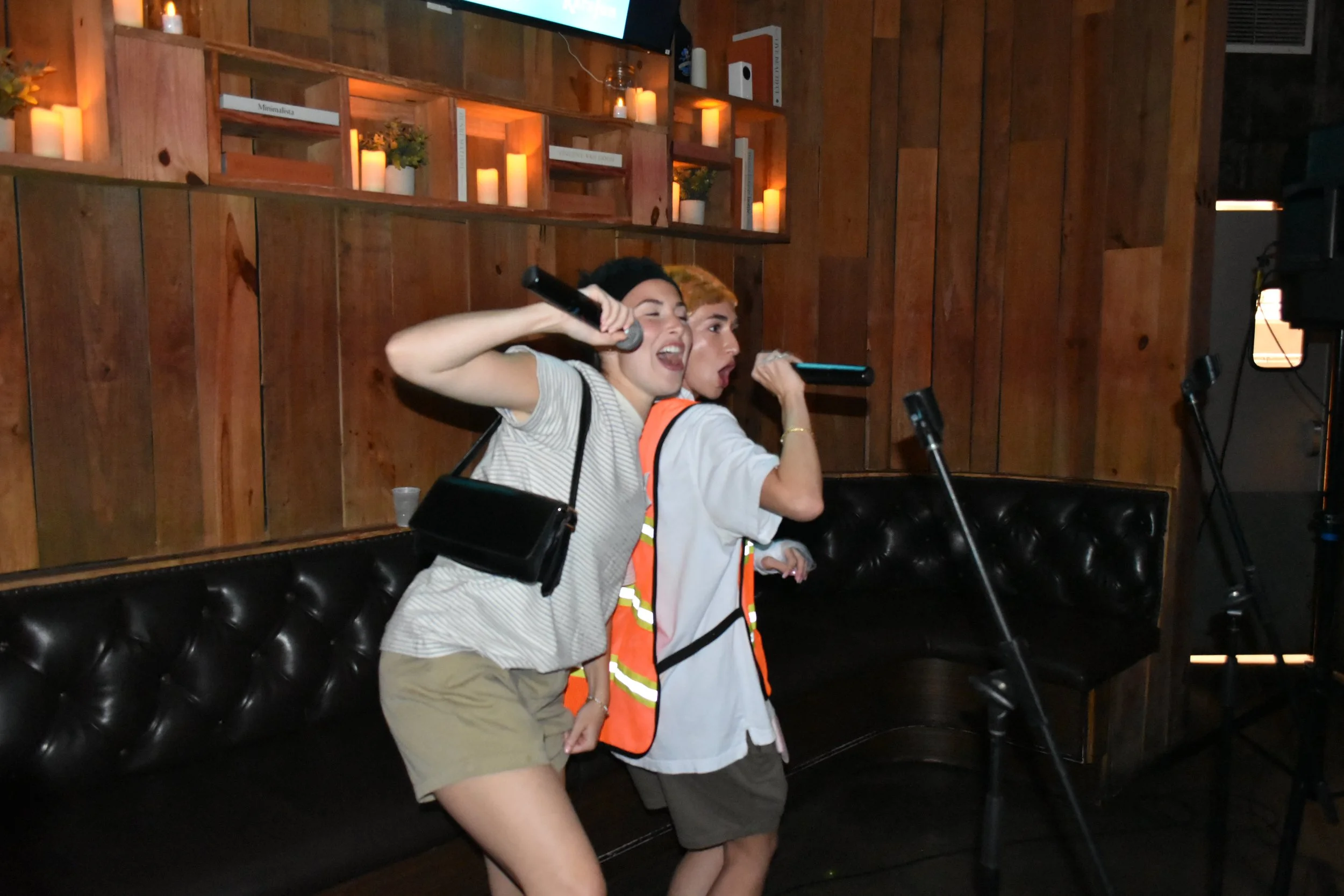 Two women singing into microphones at a karaoke bar or karaoke night, with candles on shelves and wood-paneled walls in the background.