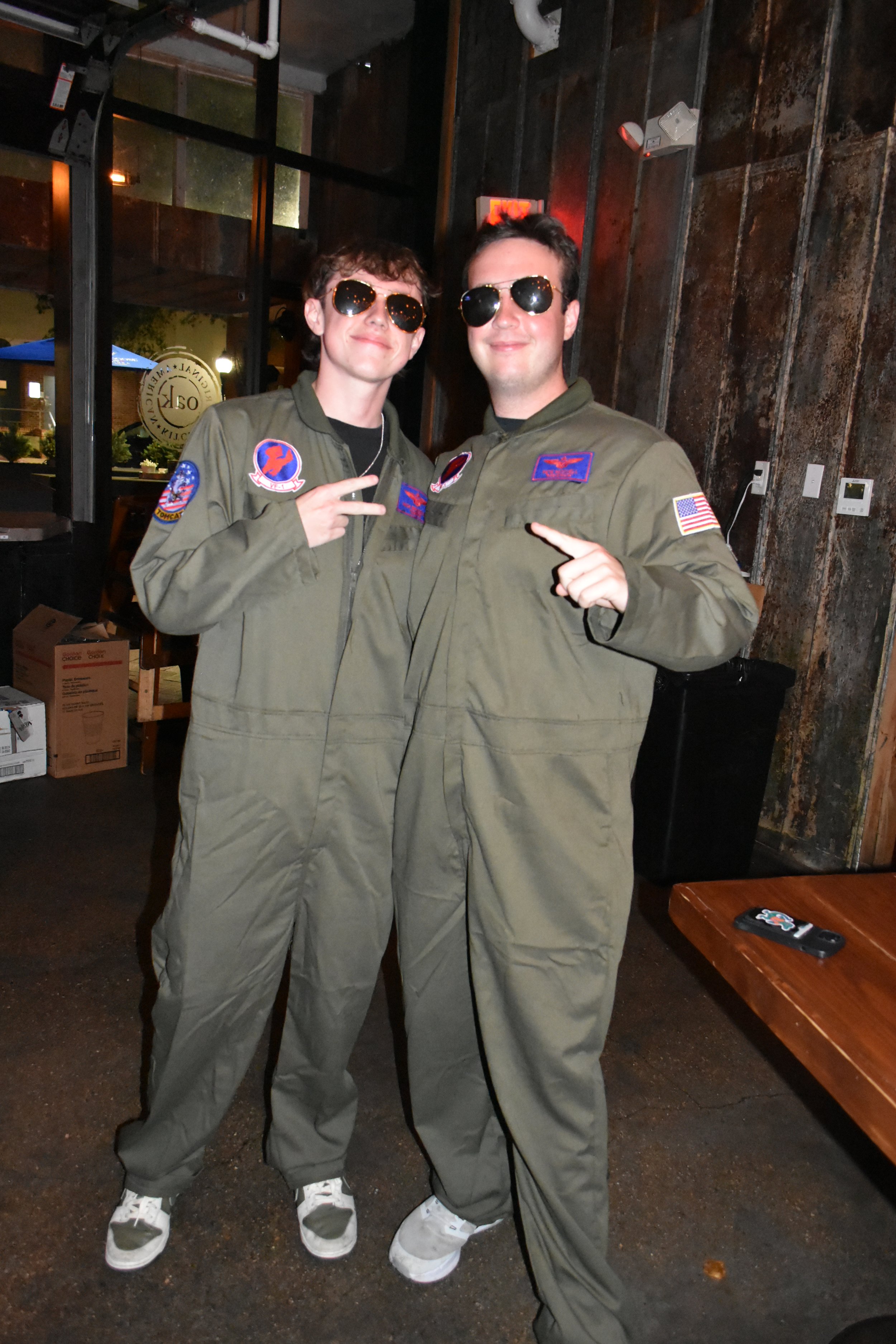 Two young men dressed in vintage NASA pilot jumpsuits, standing close together, wearing sunglasses, inside a rustic-style restaurant or cafe with wooden and brick walls.