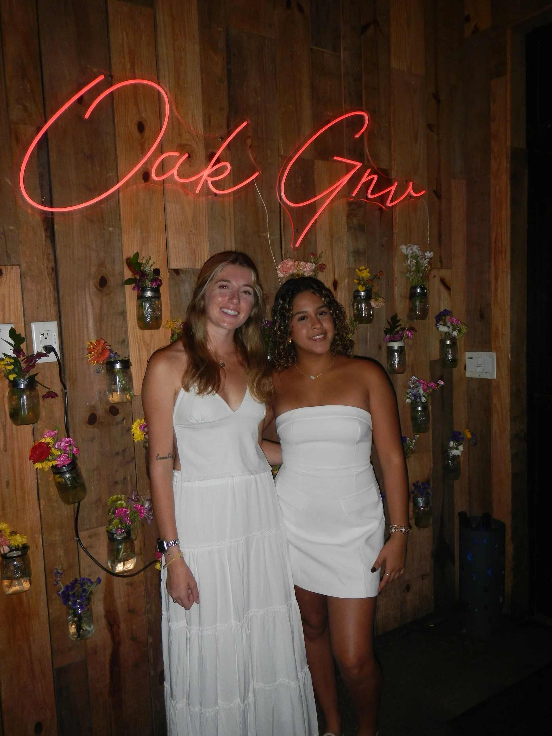 Two women wearing white dresses standing in front of a wooden wall decorated with small jars of colorful flowers and a neon sign that reads 'Jack Gnu'.