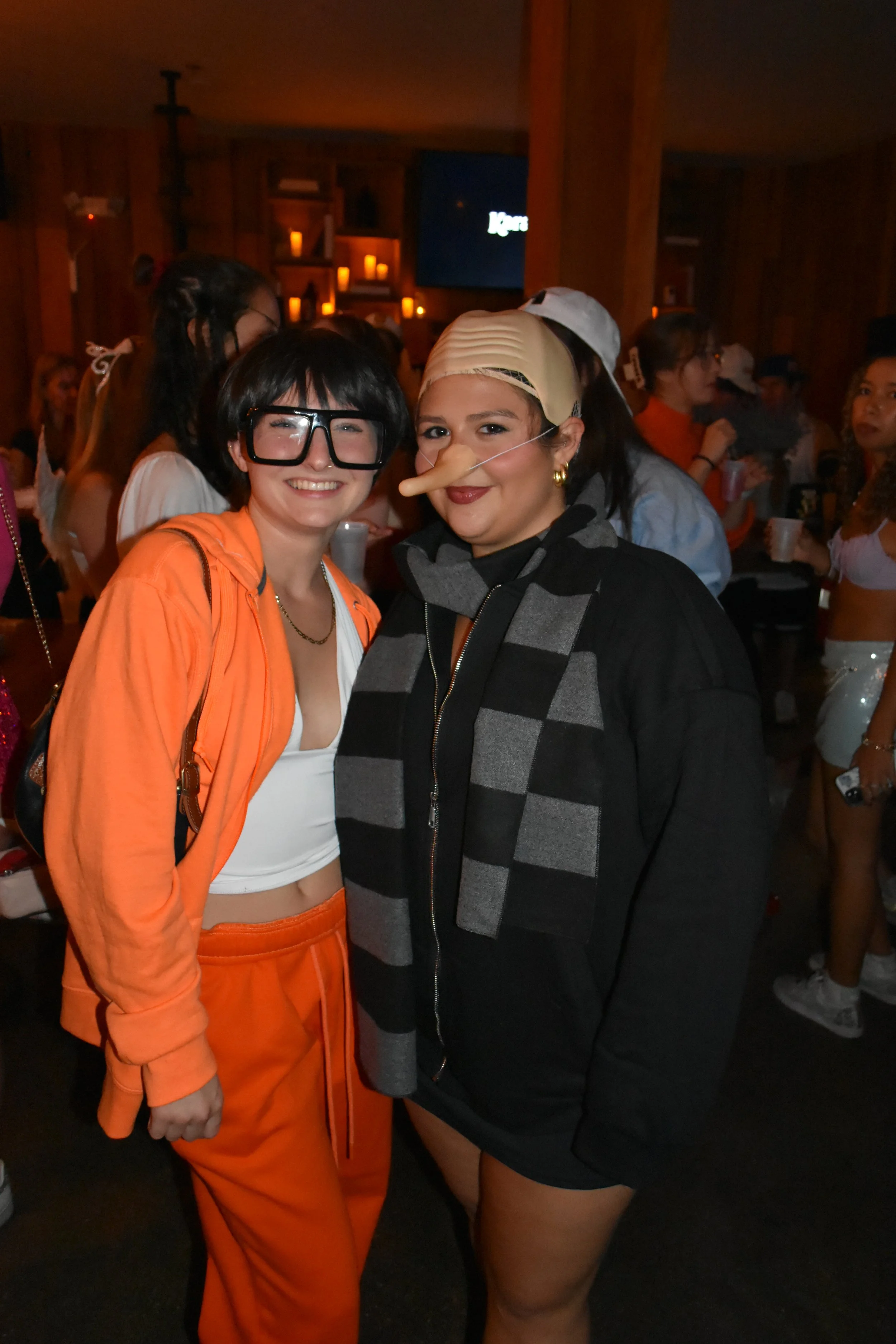 Two women in costumes at a party, one dressed as a nerd with oversized glasses and orange outfit, the other as a character with a nose and an eye patch, in a lively room with many other people in costumes.
