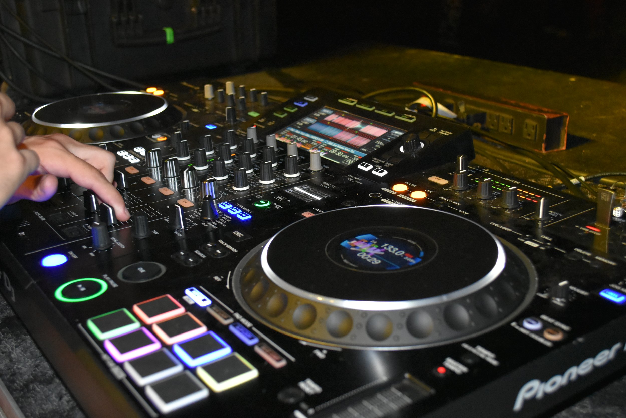 Close-up of DJ equipment with colorful lights, knobs, and a digital screen, with a hand adjusting a control.