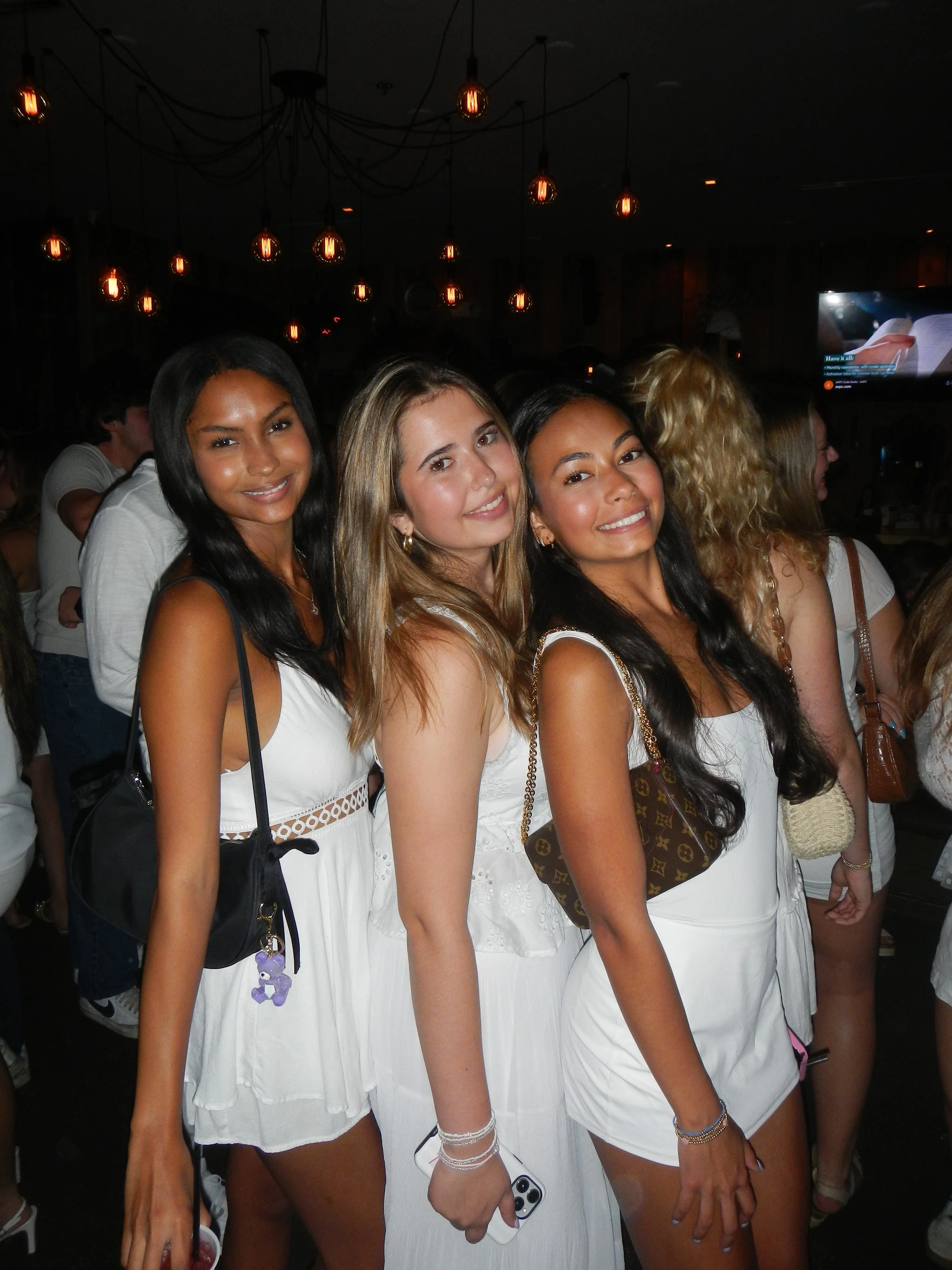 Three women smiling at a social event, dressed in white dresses, with dark hair and carrying handbags, standing close together.