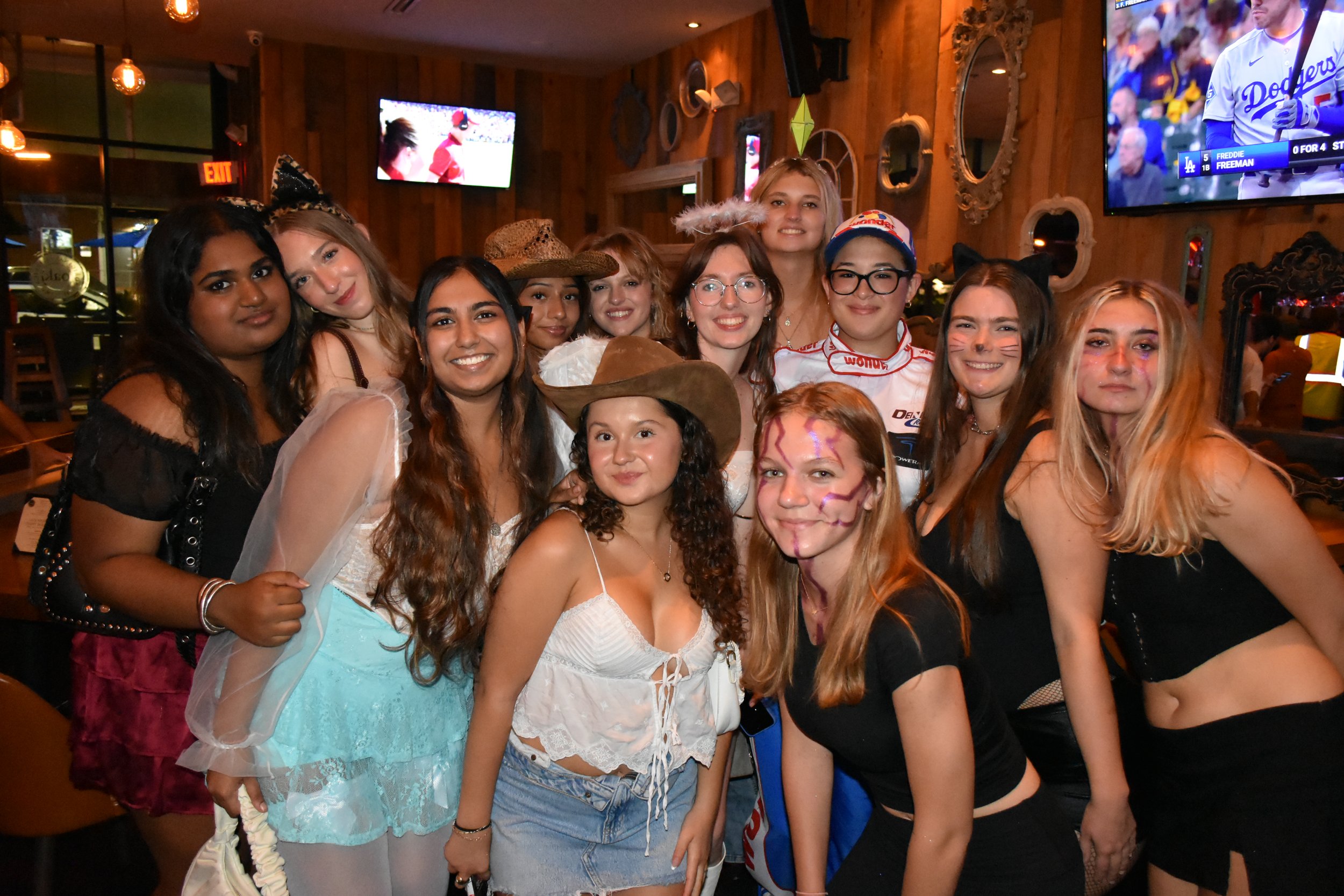 A group of young women gathered together in a bar or restaurant, dressed in costumes and casual outfits, smiling for a photo.