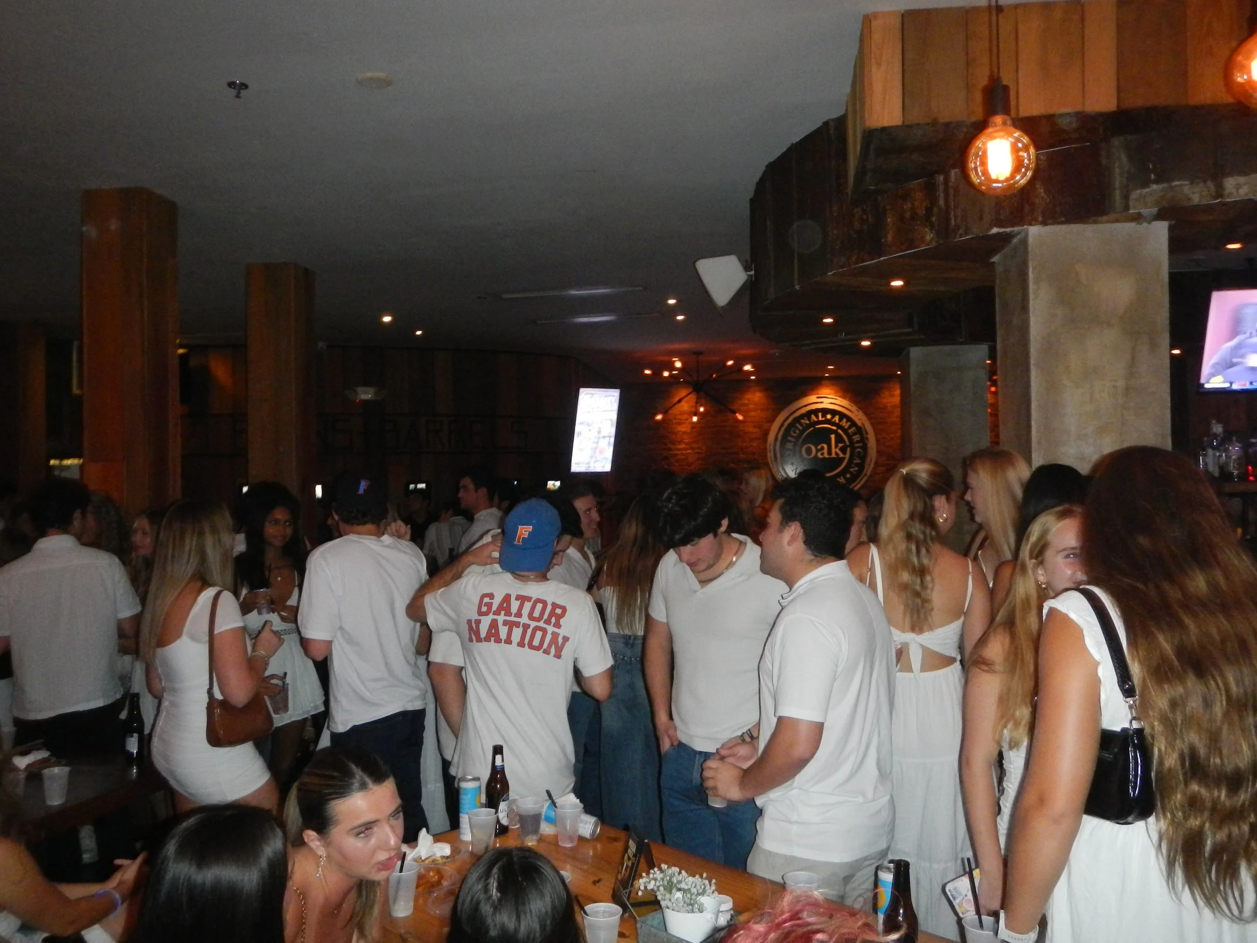 Crowd of young people at a party or bar, wearing mostly white clothing, with some holding drinks, in a dimly lit indoor setting with wooden decor.