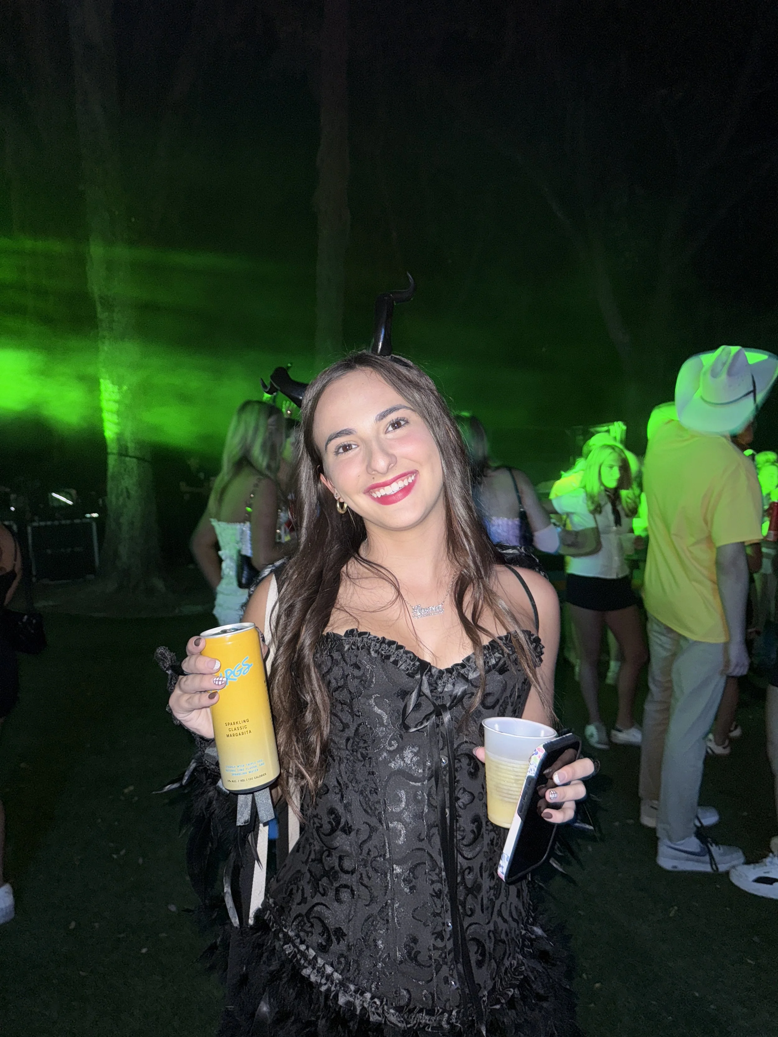 A young woman smiling at a party or concert, wearing a black dress with feather details, holding a yellow drink can and a coffee cup, wearing black devil horns headband, with green stage lights and other attendees in the background.