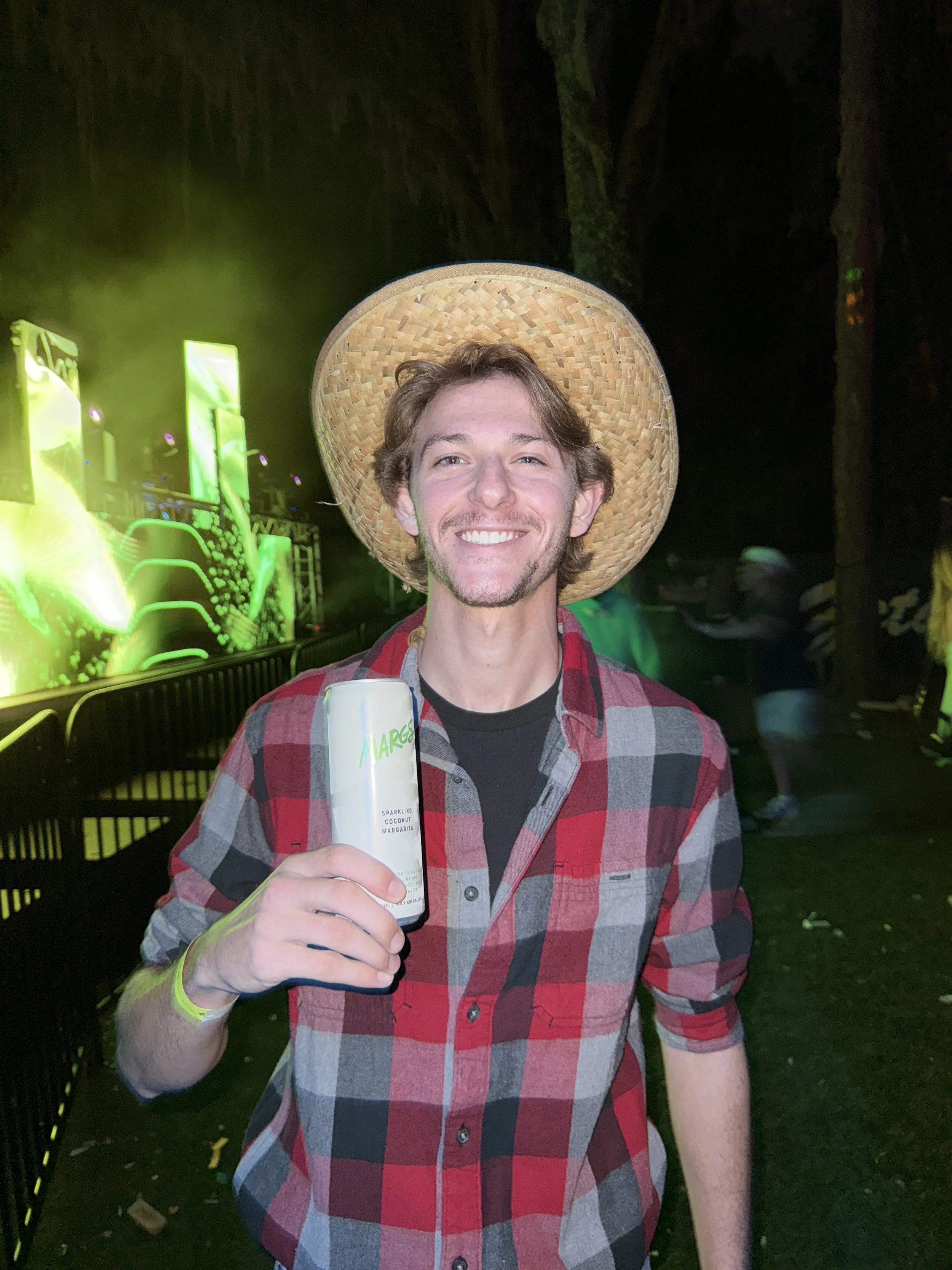 A man wearing a straw hat and a red and gray checkered shirt, smiling with a drink in hand, at an outdoor event at night with bright green lights and other people in the background.
