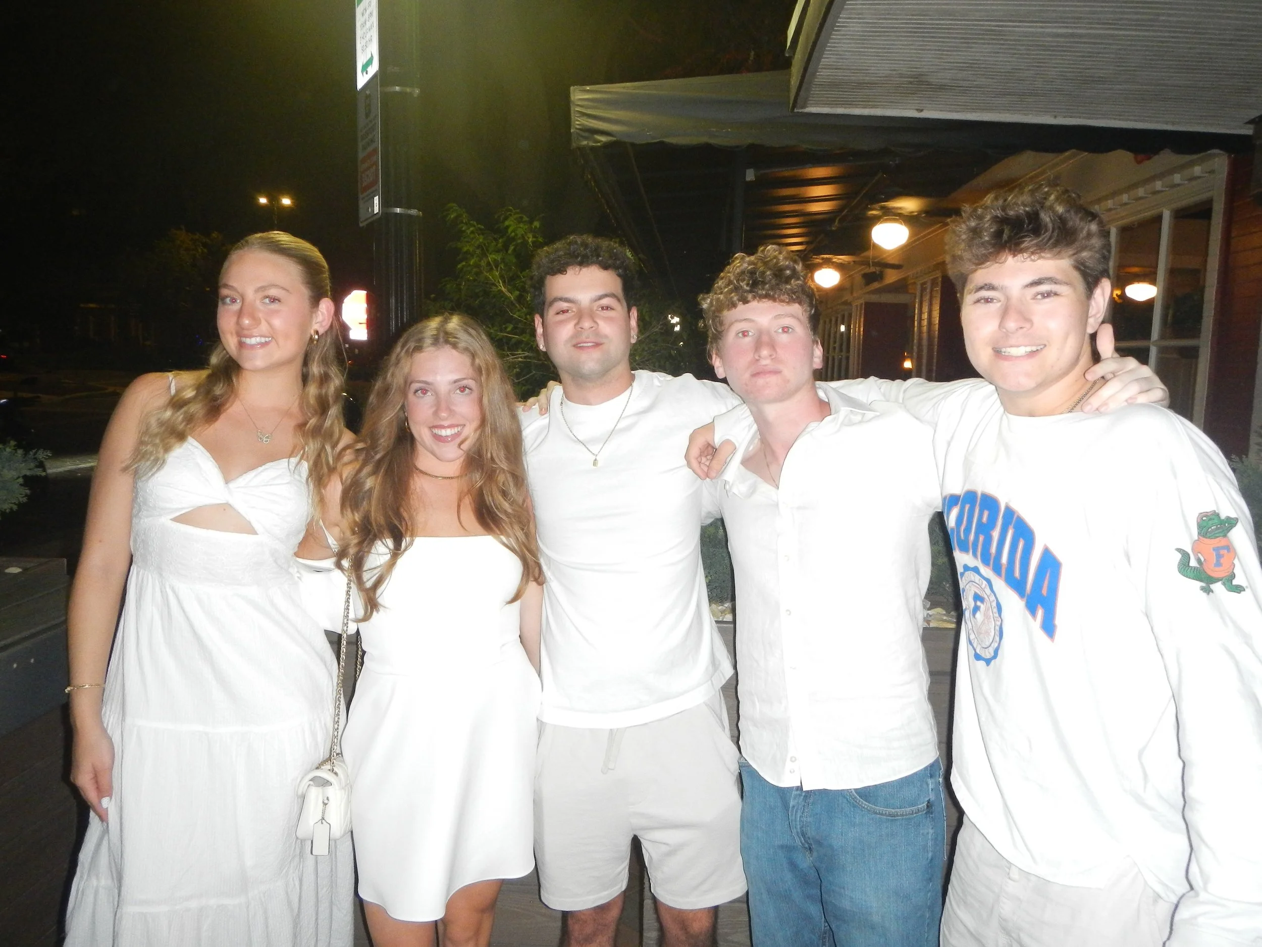 Group of five young people posing together outside at night, smiling and wearing casual summer clothing.