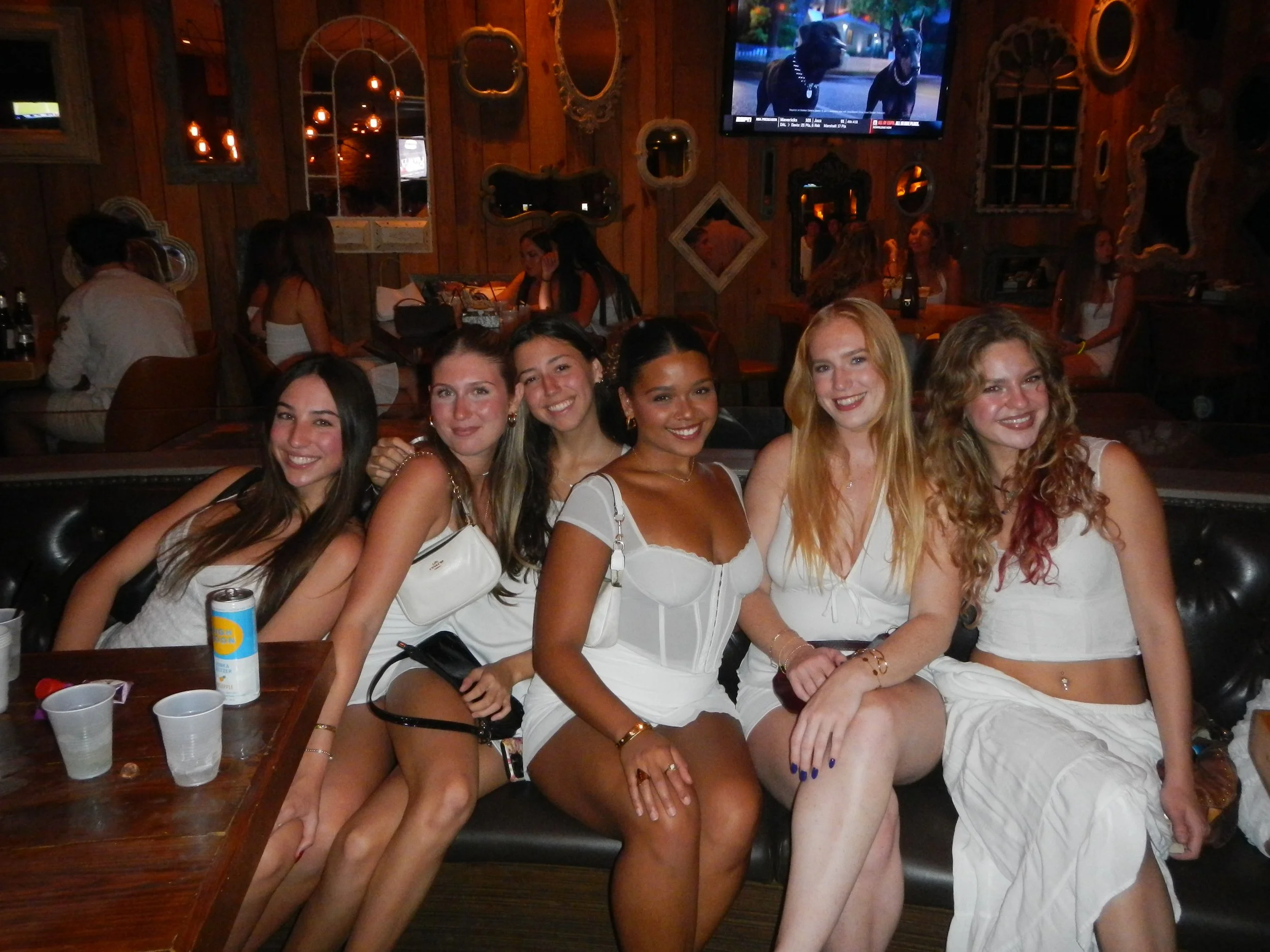 Six women sitting closely together on a black leather couch in a dimly lit bar or restaurant, smiling at the camera. The women are dressed in white or light-colored summer outfits, with some holding bags or drinks. Behind them, there are other patron