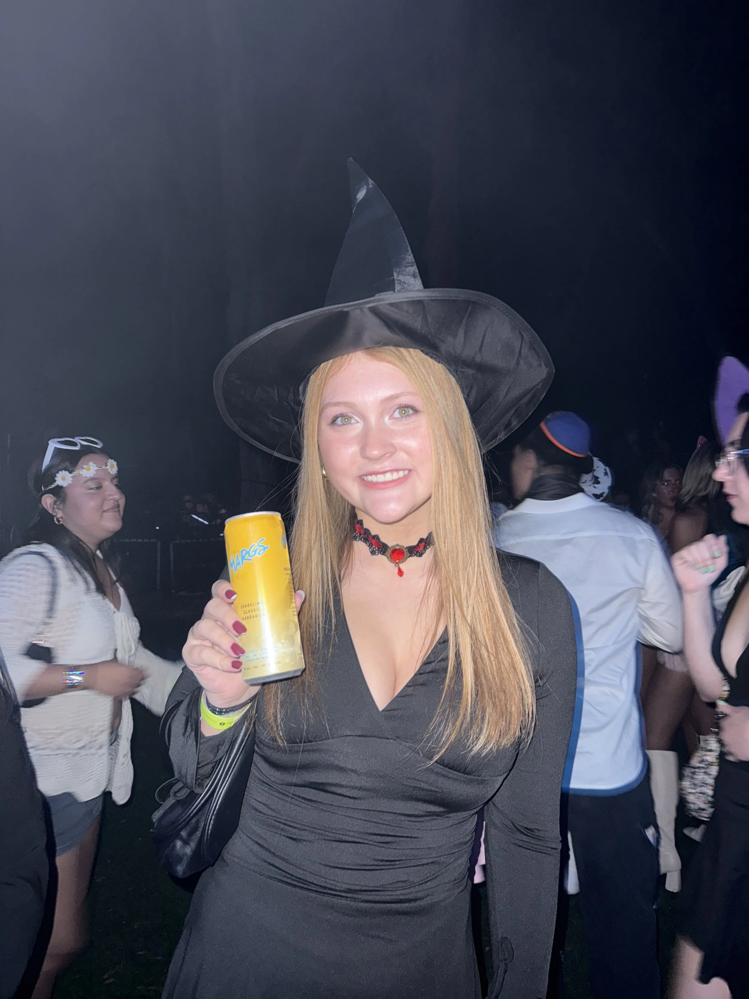 A young woman dressed as a witch at a costume party, holding a can of margarita. She has long red hair, blue eyes, and is wearing a black witch's hat and a black dress with a plunging neckline, along with a black choker necklace with a red pendant.