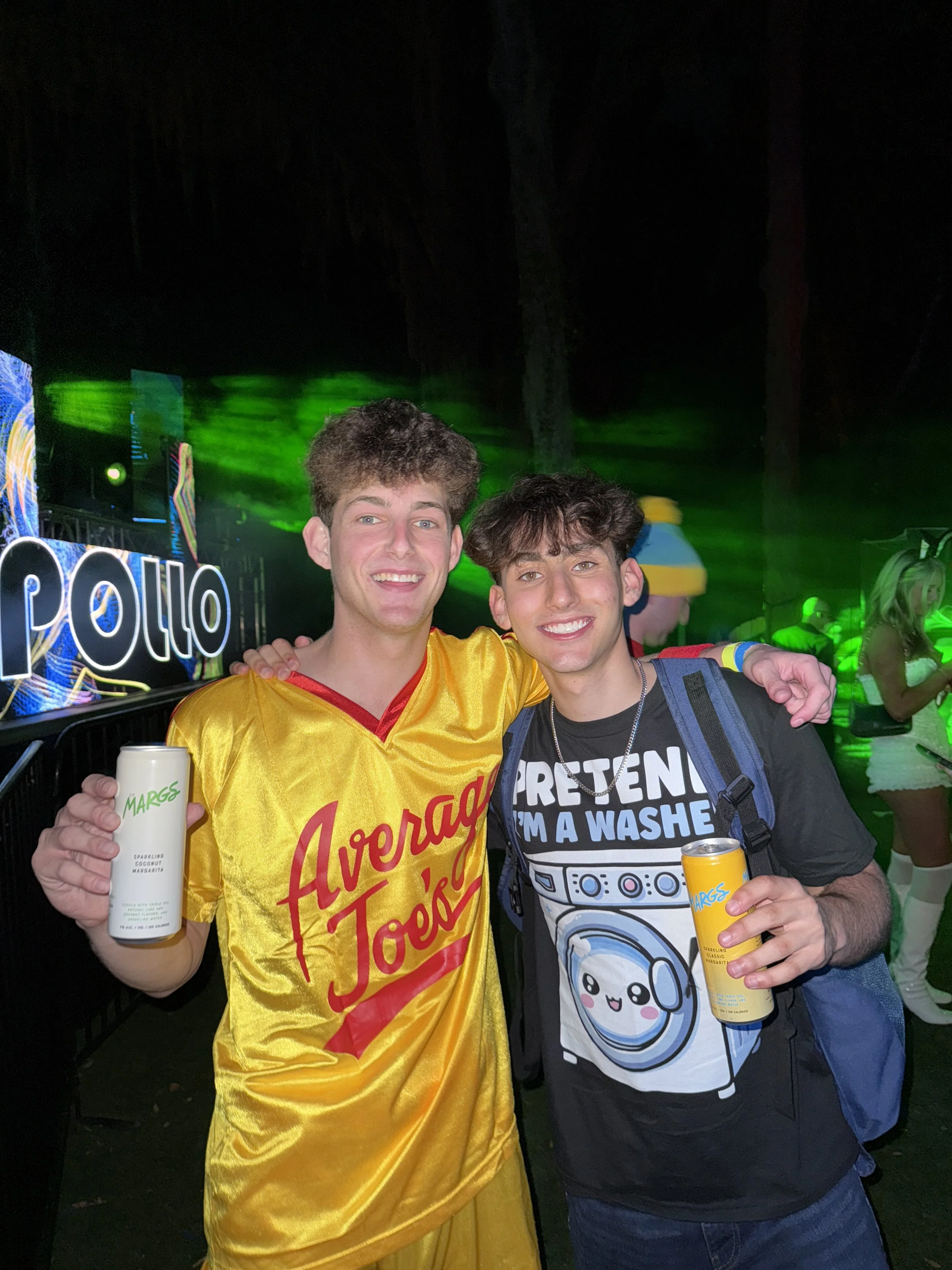 Two young men smiling and holding beverages at a lively event with green laser lights; one in a yellow basketball jersey and the other in a black graphic t-shirt with a backpack.